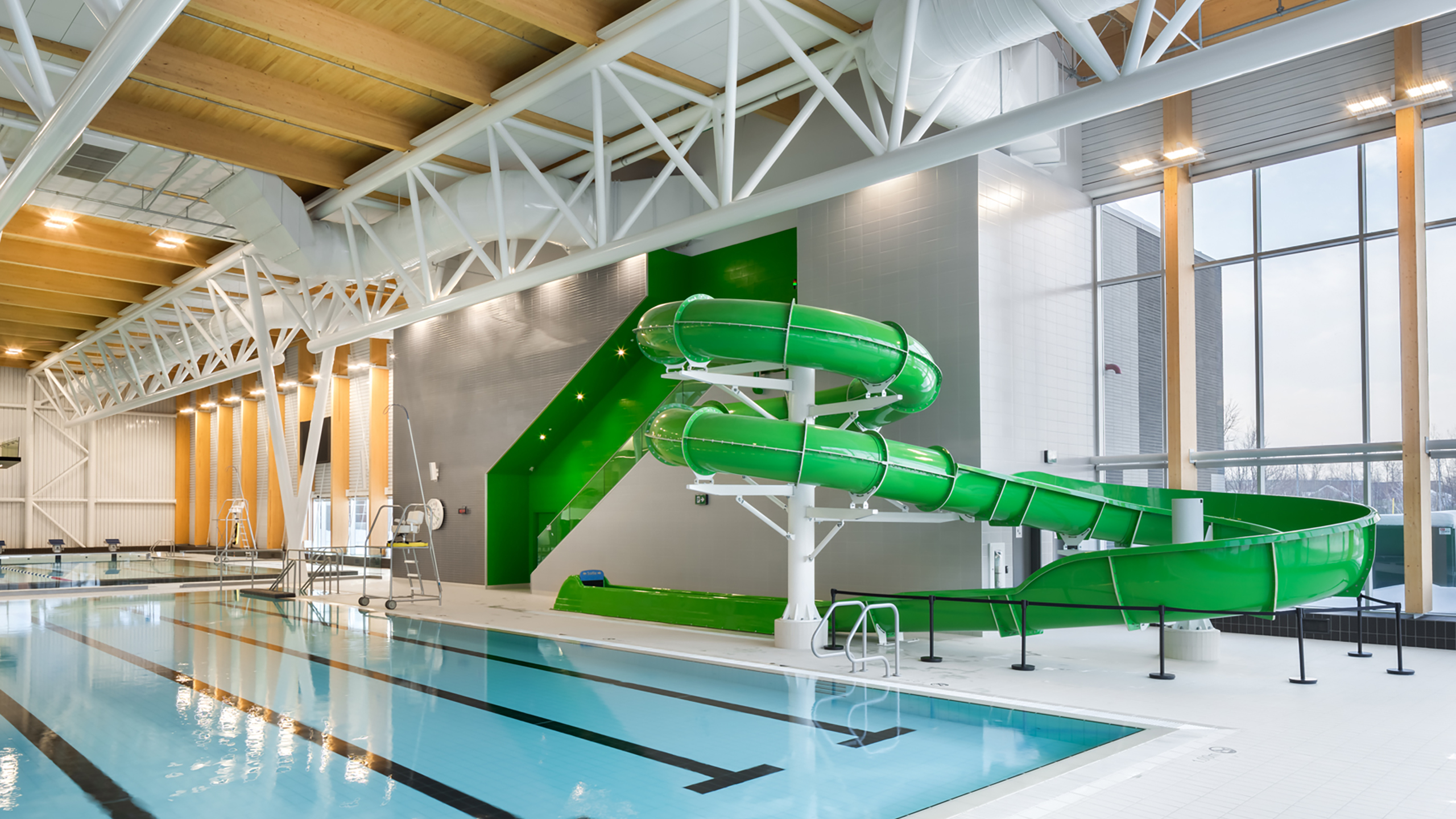 Un espace piscine intérieure au design moderne, avec un toboggan aquatique vert vif et sinueux. La piscine est entourée de carreaux de couleur claire et de grandes fenêtres laissant entrer la lumière naturelle. Dans le cadre de la construction d'un bâtiment multifonctionnel, il y a un haut plafond avec des poutres structurelles blanches et des accents en bois au-dessus.