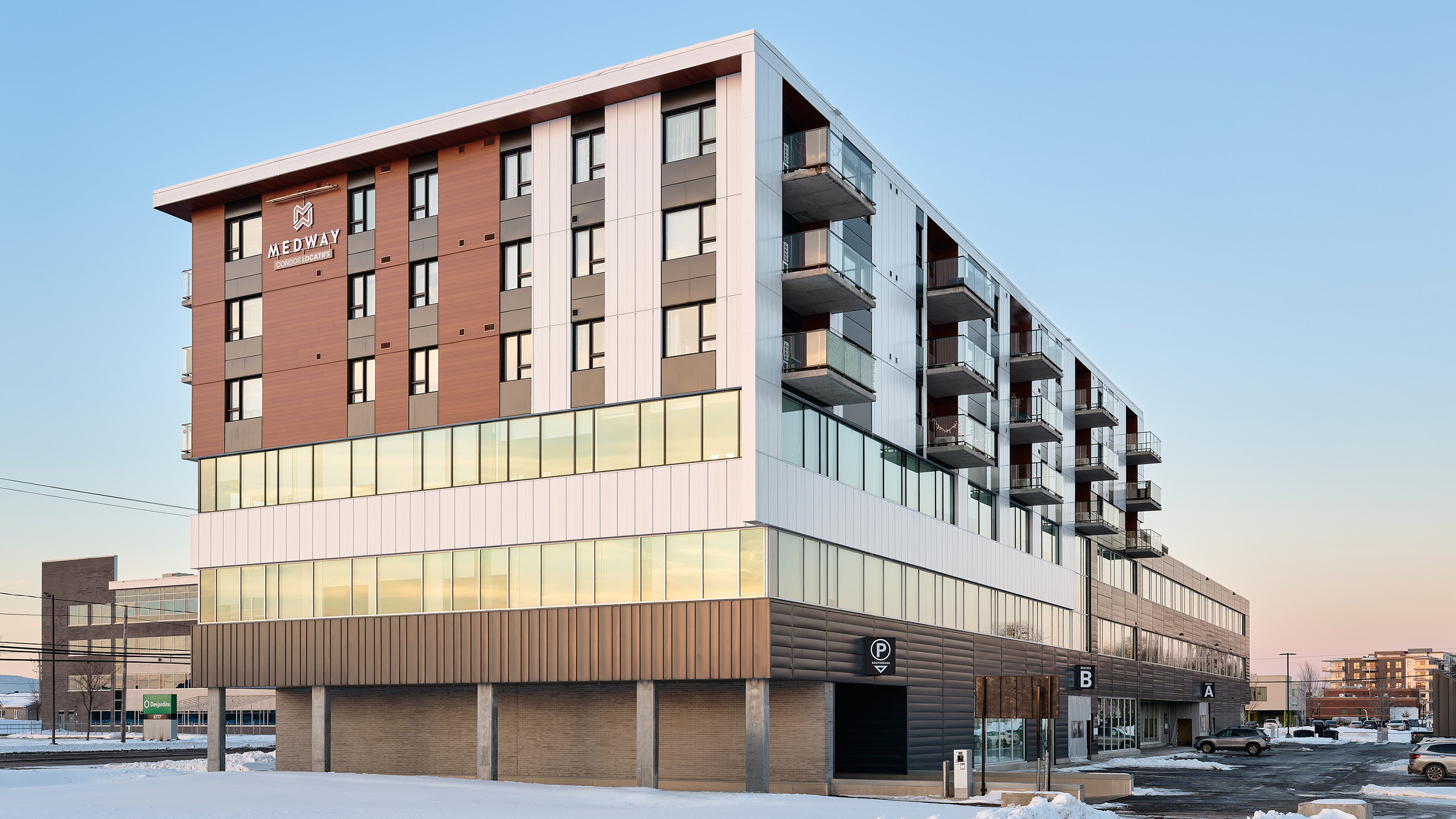 Un bâtiment moderne à plusieurs étages avec un mélange de panneaux extérieurs bruns, blancs et gris. La structure possède de nombreux balcons et de grandes fenêtres. Le rez-de-chaussée abrite des espaces commerciaux avec des enseignes éclairées. De la neige est visible sur le sol et les environs, preuve d'un agrandissement majeur en cours.