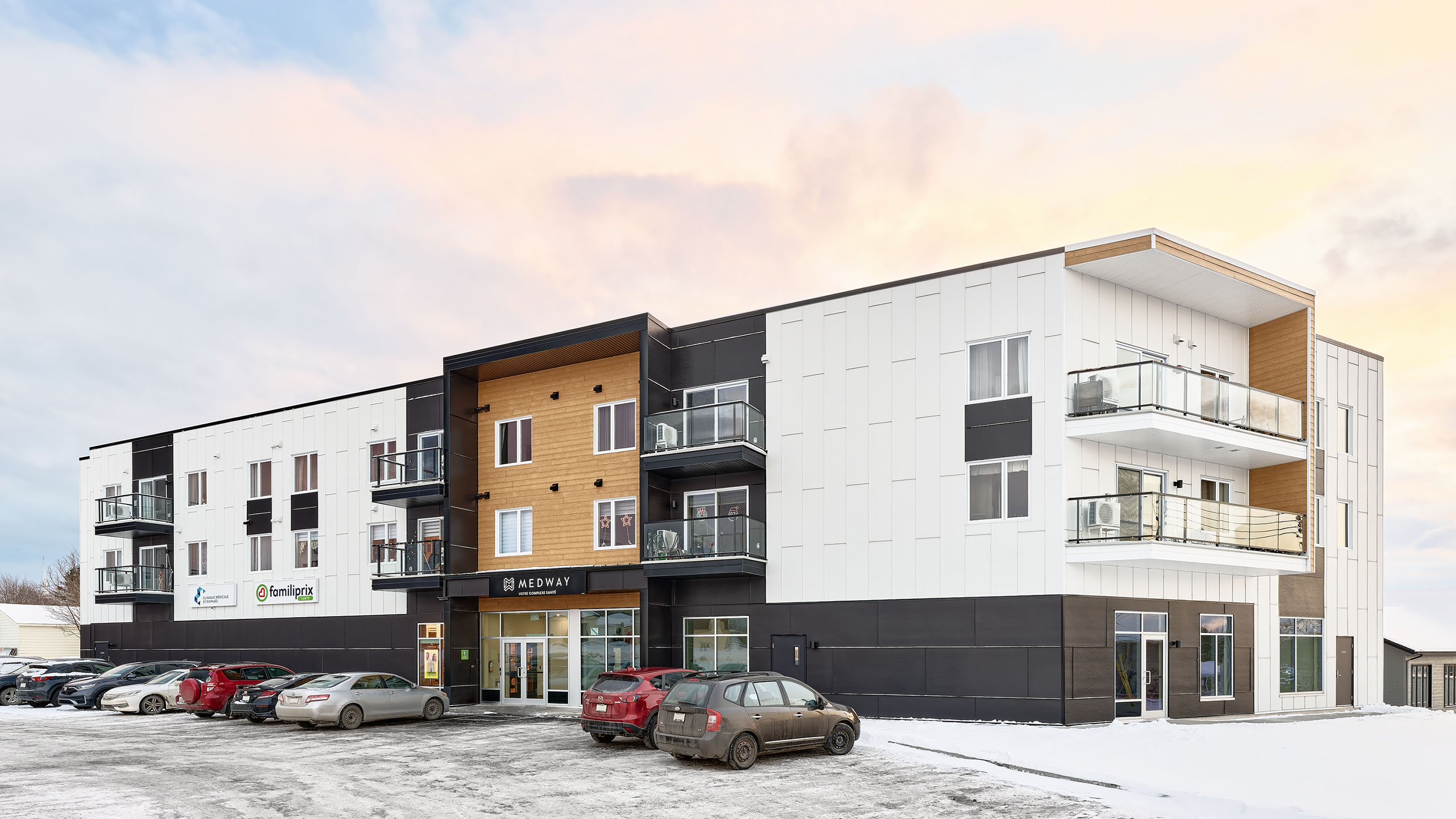 Un bâtiment moderne de trois étages avec un mélange de finitions extérieures noires, blanches et boisées met en valeur la récente construction d'un nouveau bâtiment. Des balcons ornent les deuxième et troisième étages. Plusieurs voitures sont garées devant sur un sol enneigé. L'entrée est située au centre et bien éclairée.
