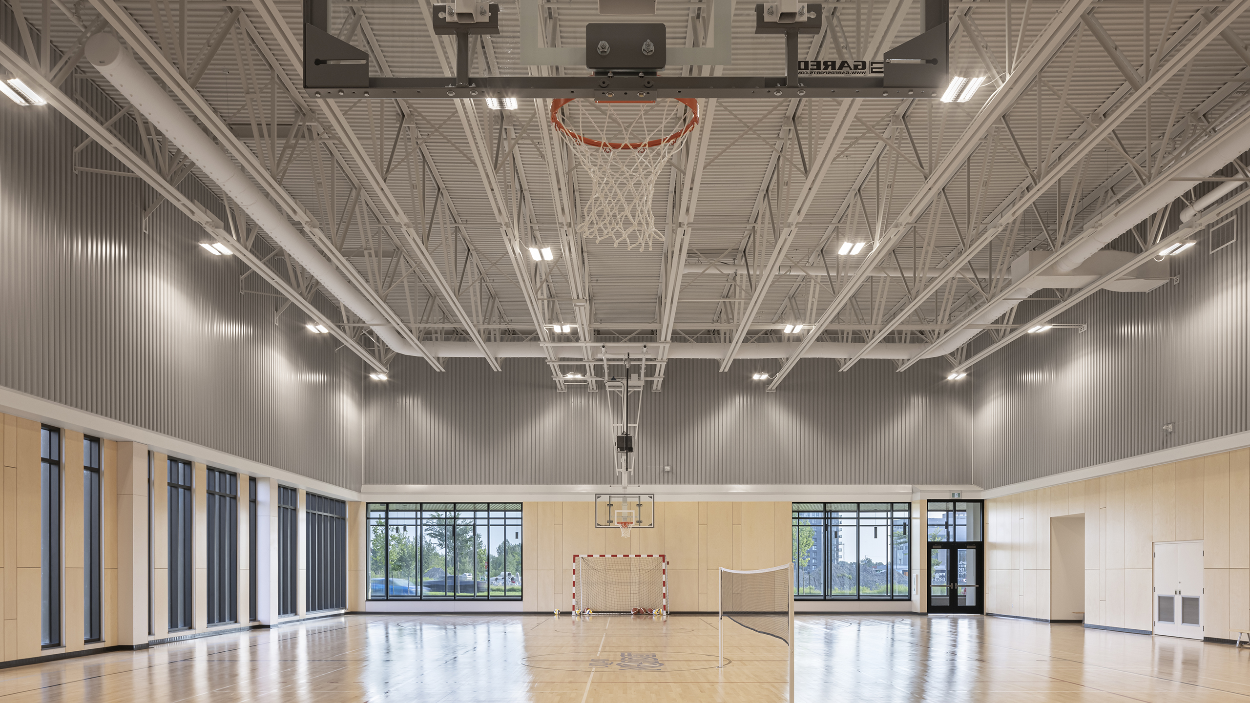 Un gymnase intérieur moderne avec de hauts plafonds, comprenant un panier de basket-ball, un filet de volley-ball et un but de soccer contre le mur du fond. De grandes fenêtres sur la gauche permettent à la lumière naturelle d'inonder le plancher en bois poli. Une partie de la construction d'un complexe sportif, des portes sont également visibles en arrière-plan.