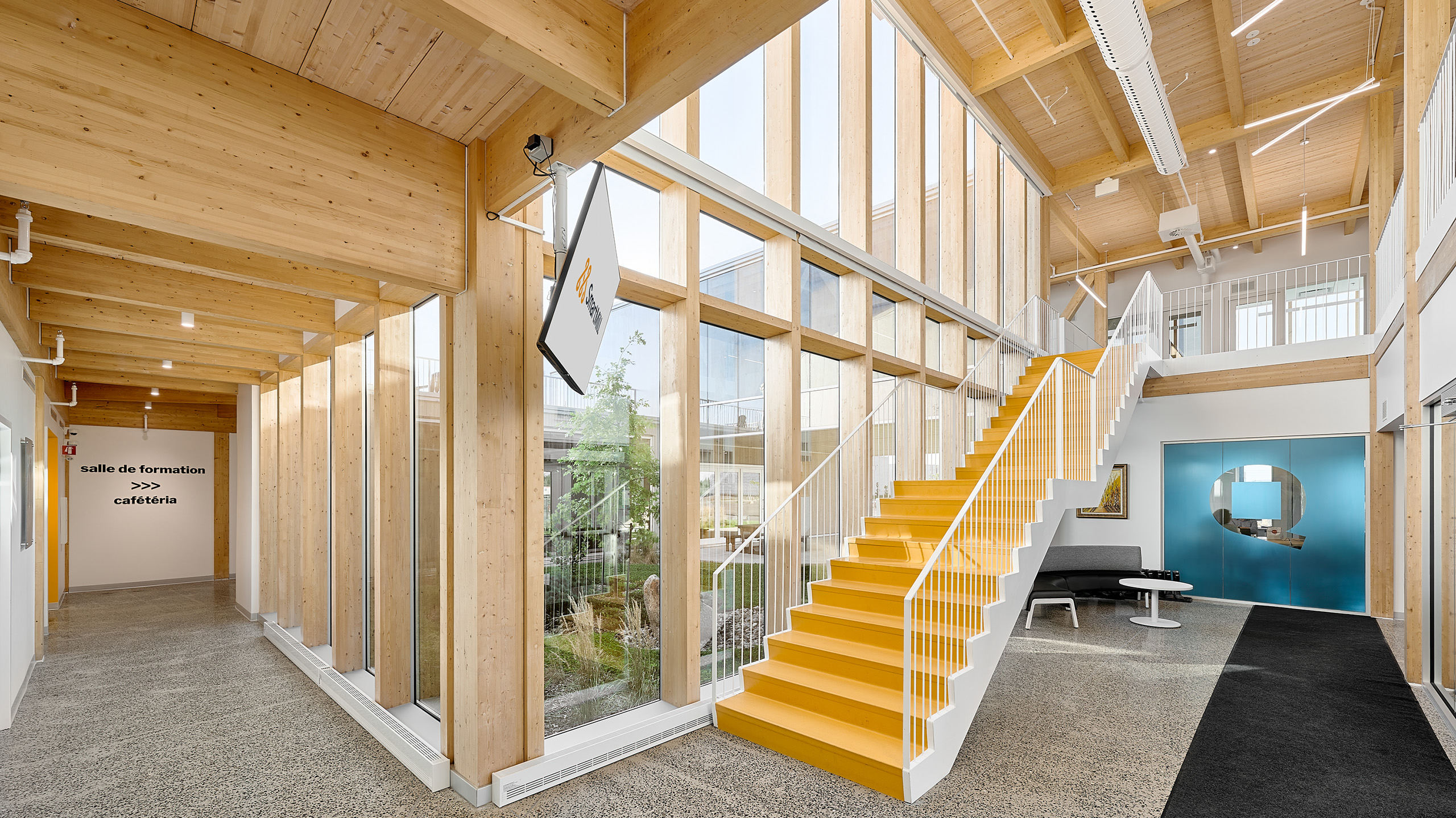 Un intérieur moderne avec de hautes fenêtres et des poutres en bois présente un escalier droit jaune vif. Il y a un couloir sur la gauche avec un panneau indiquant « salle de formation » et « cafétéria ». Dans le cadre de la construction d'un nouveau siège social, un canapé noir et une œuvre d'art bleue sont visibles en arrière-plan.
