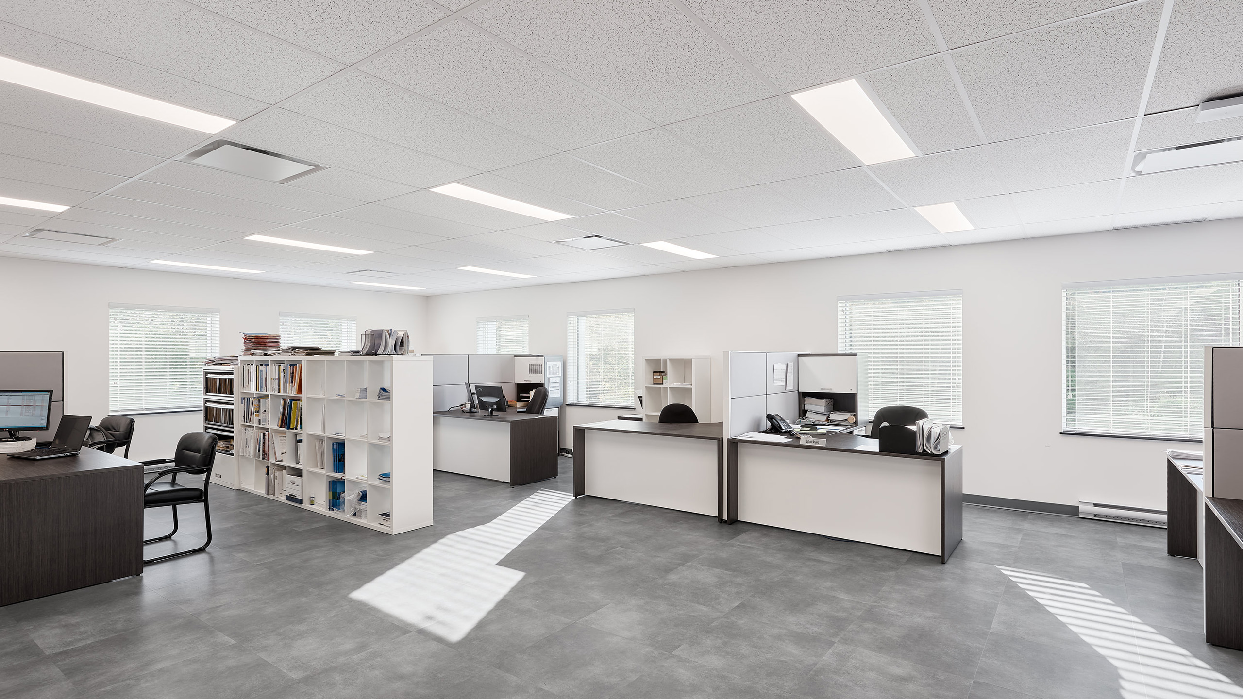 Un bureau lumineux et moderne avec plusieurs postes de travail séparés par des cloisons blanches. Chaque bureau est équipé de fournitures de bureau, d'ordinateurs et de chaises. De grandes fenêtres bordent les murs, permettant à la lumière naturelle de remplir l'espace. Dans le cadre de la construction d'un nouveau bâtiment, des étagères accueillent livres et documents.
