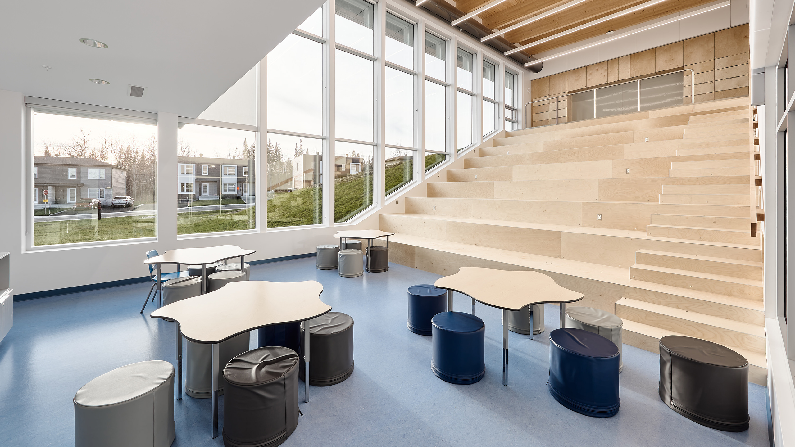 Une salle moderne et bien éclairée avec de grandes fenêtres, des sièges en bois à plusieurs niveaux et diverses tables aux formes uniques entourées de tabourets ronds et rembourrés. Le sol bleu complète la lumière naturelle provenant de la construction d'une nouvelle école à l'extérieur, offrant une vue sur un espace extérieur gazonné et des maisons.