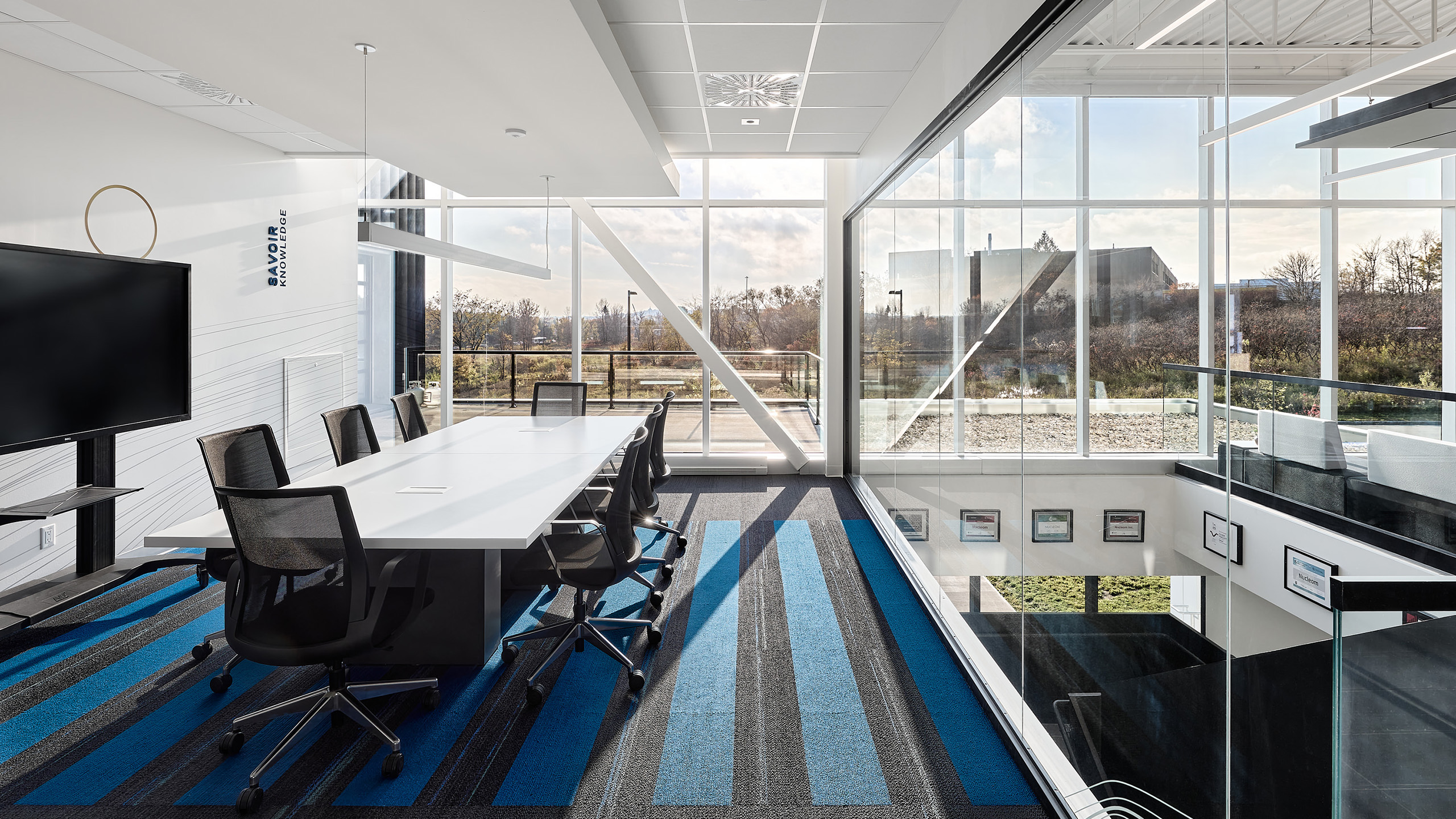 Une salle de conférence moderne avec une longue table blanche, entourée de chaises de bureau sombres sur un tapis rayé bleu et gris. Une extrémité de la pièce dispose de grandes fenêtres avec vue sur un espace extérieur, reflétant la construction d'un nouveau siège social. Un téléviseur fixé au mur orne la gauche tandis que des tableaux encadrés décorent la droite.