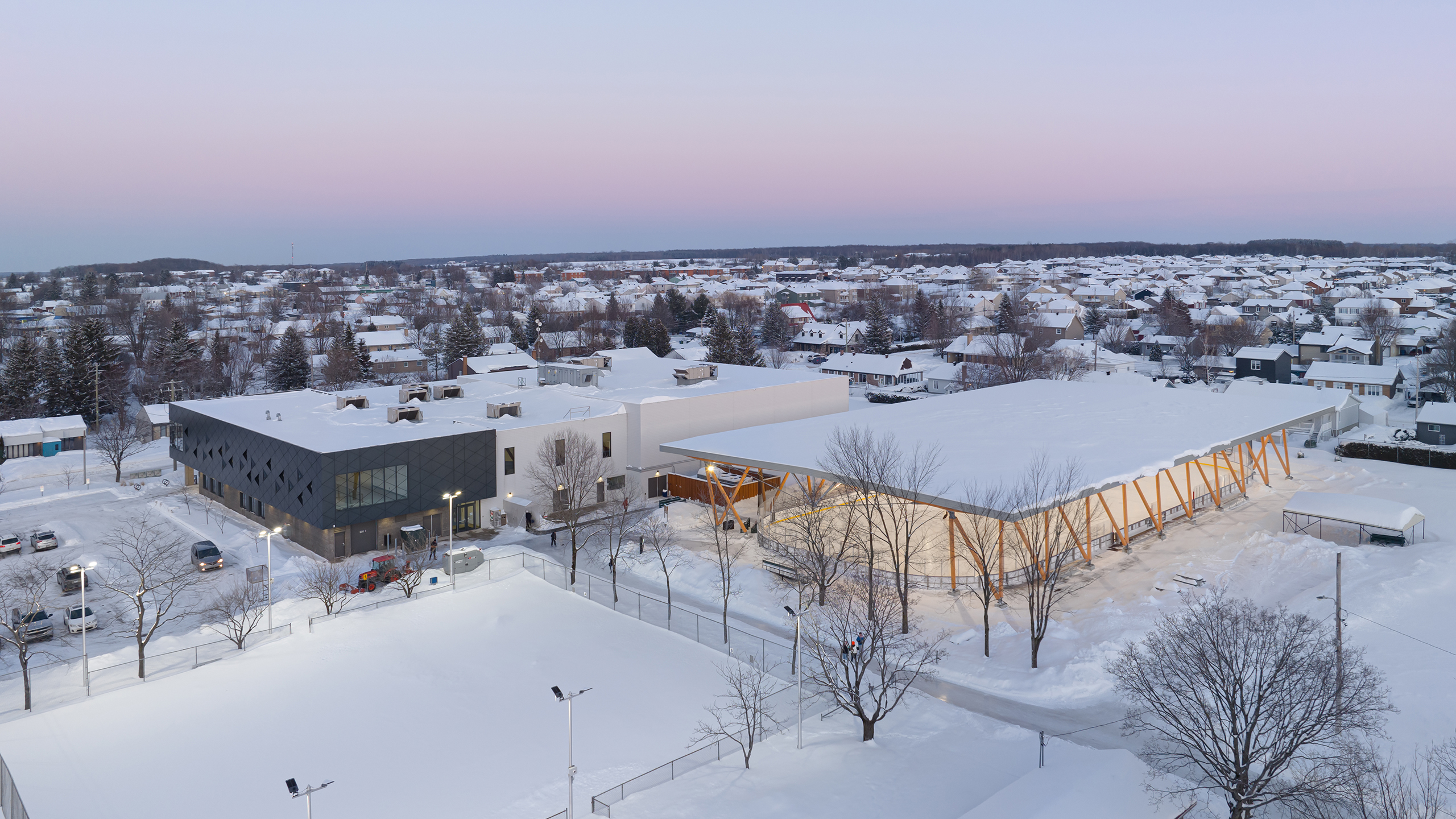 Vue aérienne d'un paysage urbain enneigé en hiver, avec un grand complexe de bâtiments modernes aux extérieurs à la fois sombres et clairs, témoignage d'une conception et d'une construction magistrales. La zone comprend des installations sportives, entourées de maisons résidentielles et d'arbres recouverts de neige sous un ciel pastel.