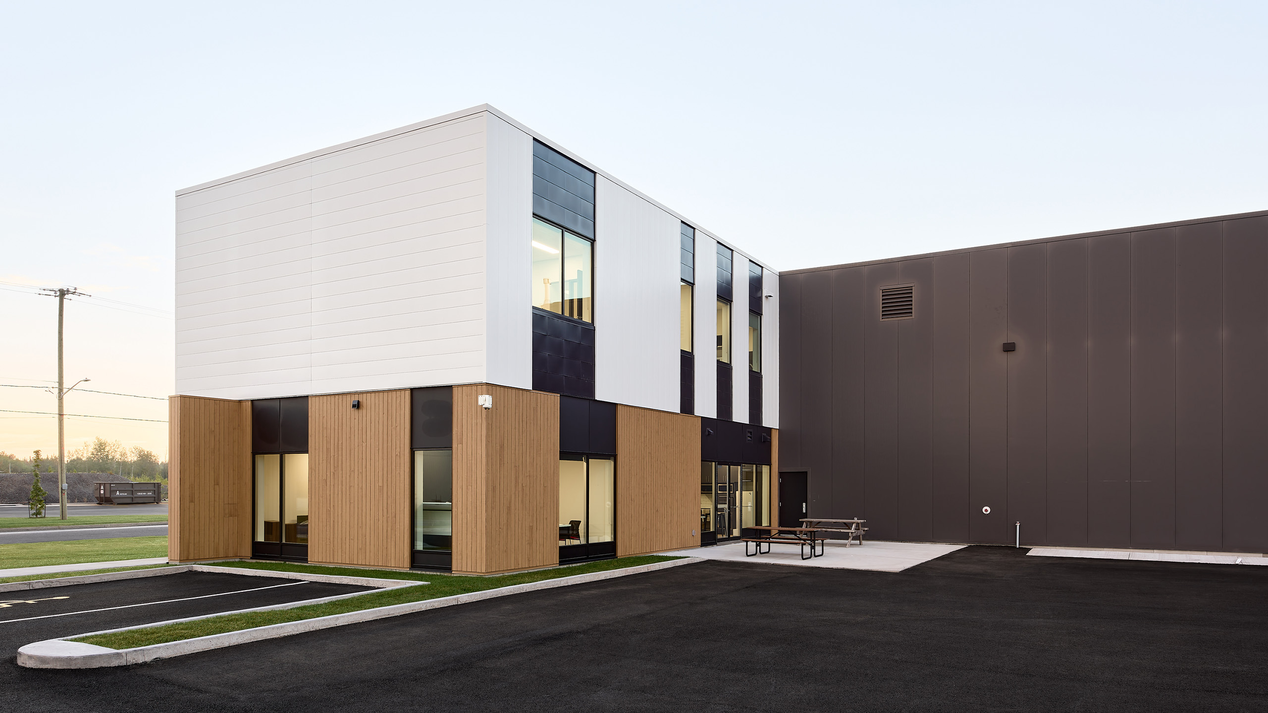 La construction du nouveau siège social présente un design moderne à deux étages avec un mélange de panneaux extérieurs blancs, noirs et en bois. Il est doté de grandes fenêtres et d'un toit plat. Les environs comprennent un parking pavé, une pelouse et une table de pique-nique près de l'entrée sous le ciel bleu clair.