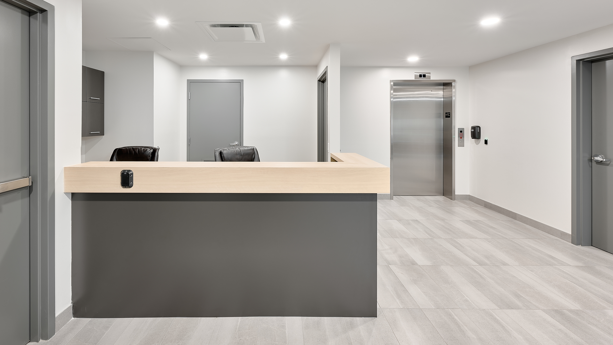Un espace de réception moderne et minimaliste dans la construction d'un nouveau bâtiment comprend un comptoir en bois avec deux chaises noires derrière lui et des portes grises en arrière-plan. À droite, il y a un ascenseur fermé avec un panneau de commande à côté. Le sol est carrelé en gris clair et les murs sont blancs.