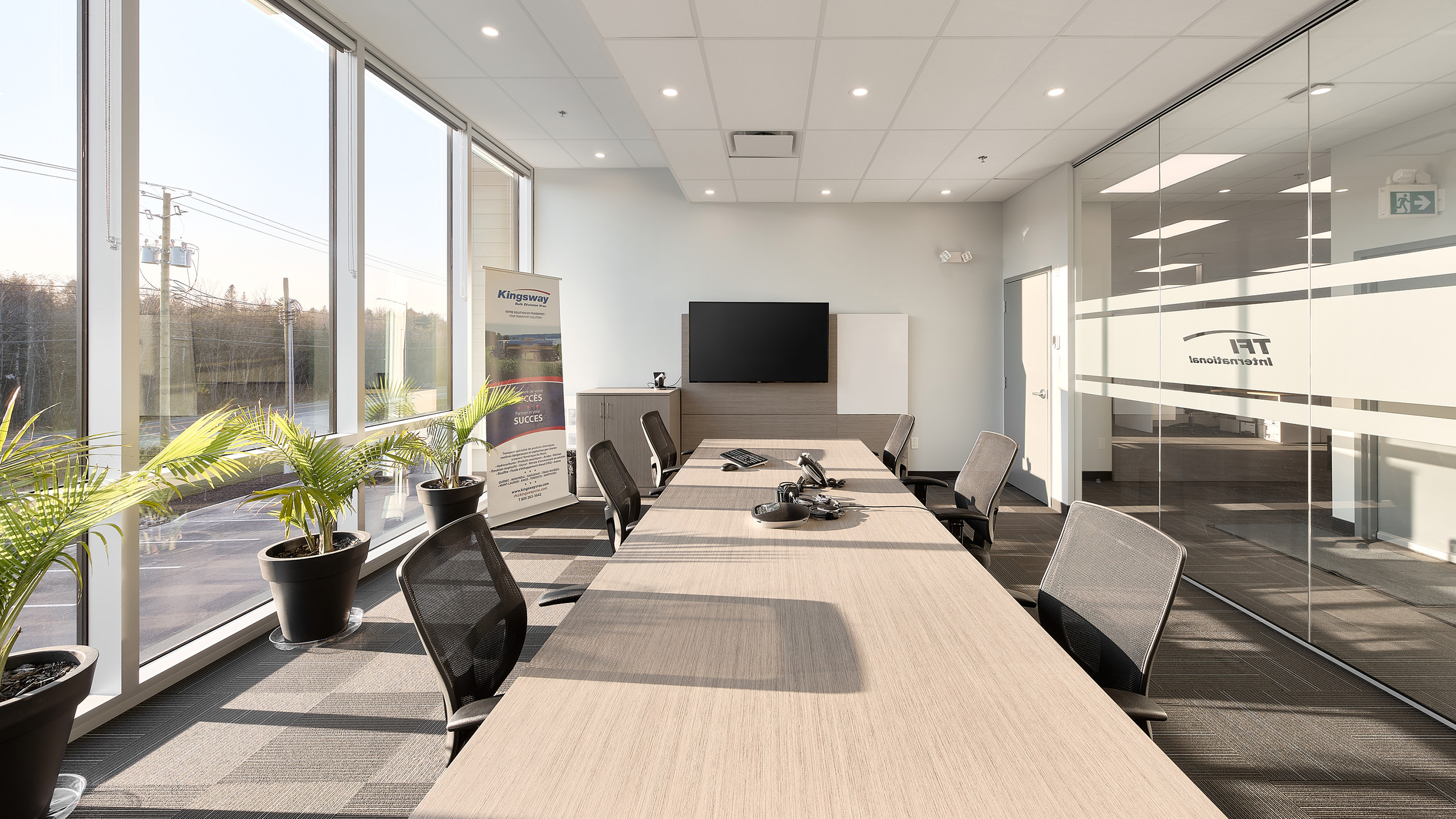 Une salle de conférence moderne avec une longue table en bois entourée de chaises de bureau noires. La salle dispose de grandes fenêtres, de plusieurs plantes en pot, d'une télévision à écran plat sur le mur et d'une bannière dans le coin mettant en valeur la construction d'un nouveau garage. Les murs en verre affichent le logo du bureau en givrage.