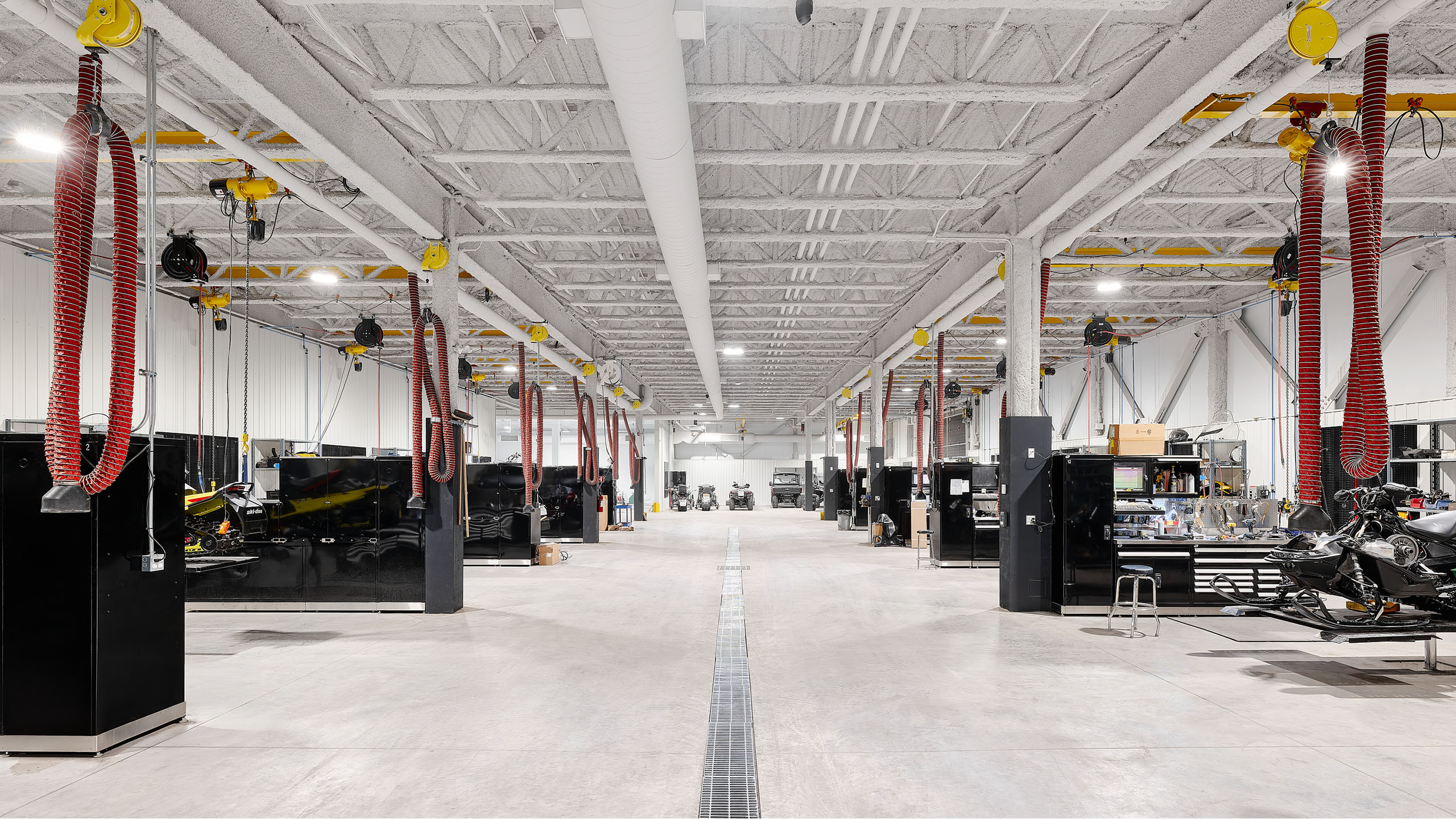 Un atelier de réparation automobile spacieux et bien éclairé dans un bâtiment neuf dispose de postes de travail des deux côtés. Des tuyaux spiralés rouges pendent du plafond et divers équipements et outils sont disposés à chaque poste. Le plafond et les murs blancs créent un environnement propre et ordonné.