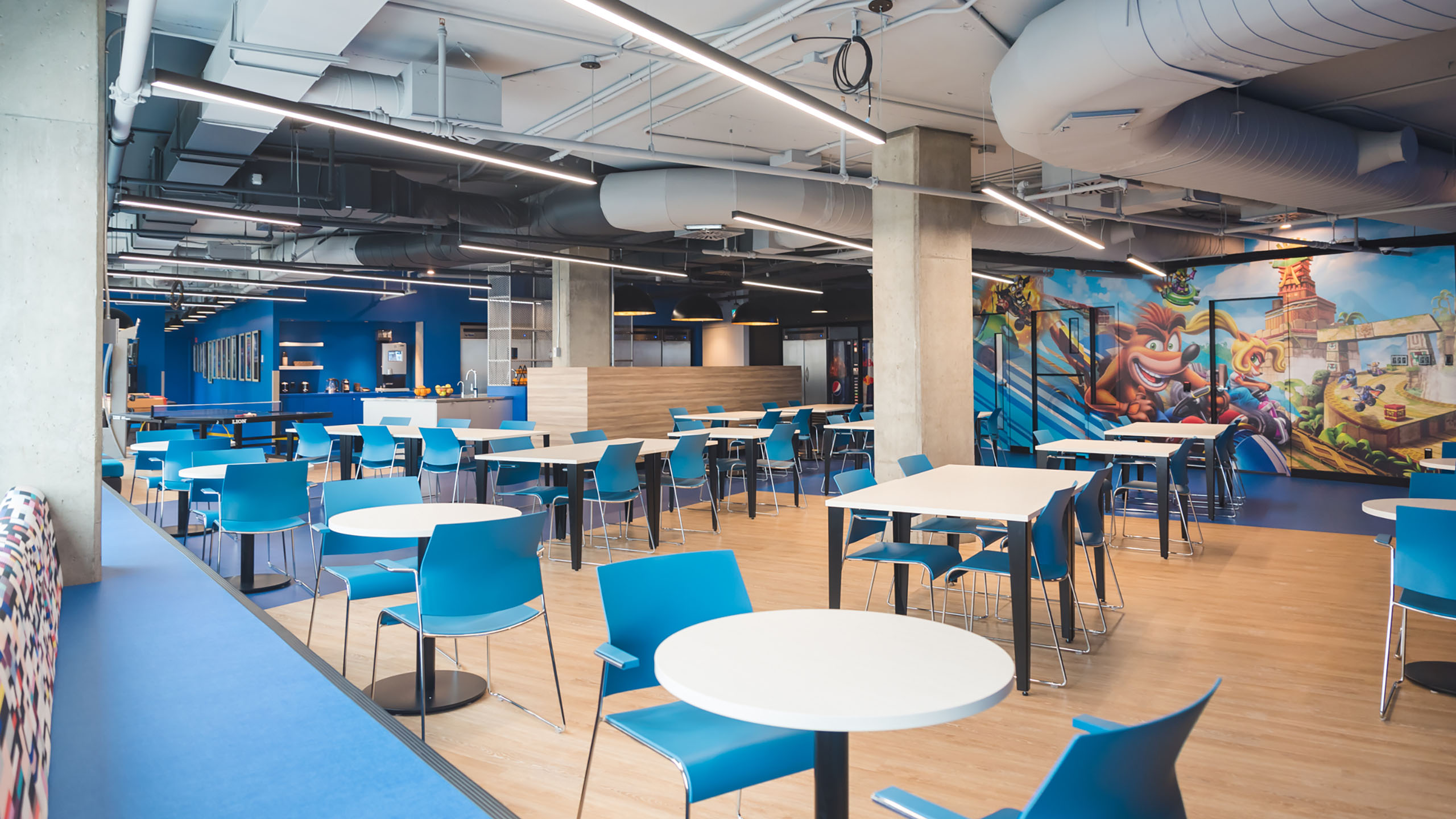 Une cafétéria lumineuse avec des chaises bleues modernes et des tables blanches, récemment rénovée grâce à la rénovation des espaces bureaux. L'espace présente des conduits apparents au plafond et une grande fresque murale colorée représentant des personnages animés. Il y a un comptoir à l'arrière avec diverses options de restauration et de boissons.