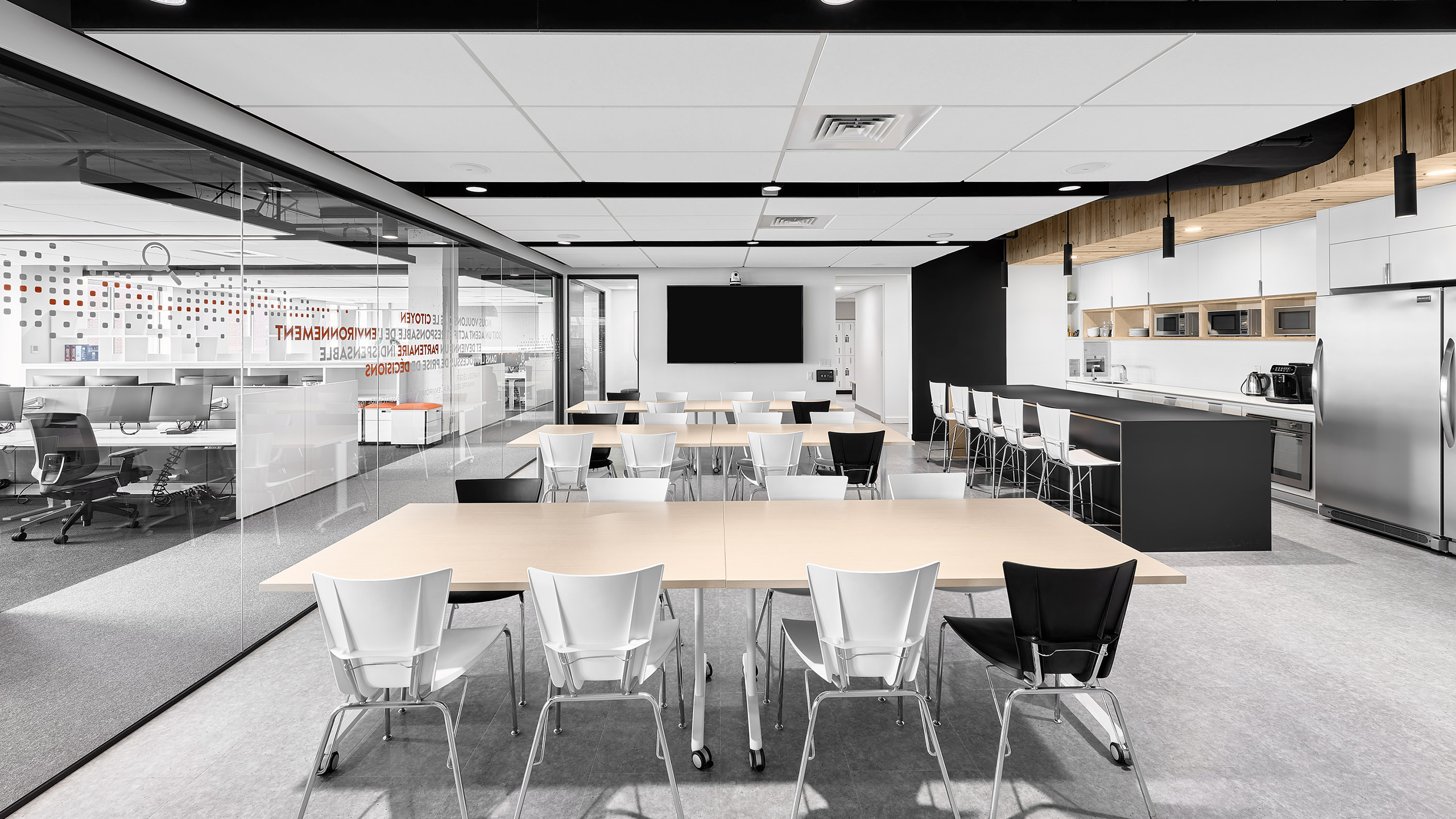 Un espace de pause de bureau moderne avec plusieurs tables en bois clair entourées de chaises blanches et noires. En arrière-plan, on trouve une cuisine avec un réfrigérateur en acier inoxydable, des micro-ondes et des armoires de rangement. Un grand écran noir est fixé au mur. Le côté gauche montre un espace de bureau adjacent visible à travers une cloison vitrée, mettant en valeur un aménagement intérieur des bureaux impeccable.