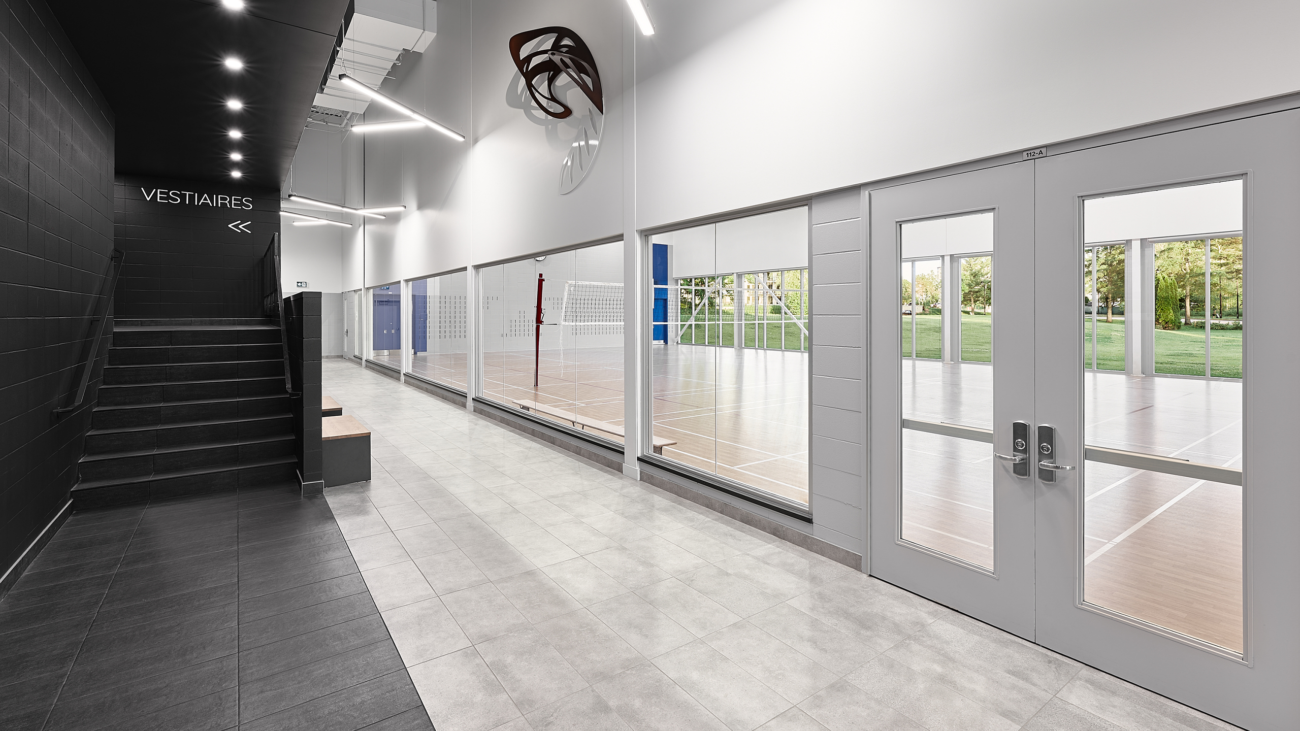 Un gymnase intérieur moderne aux parois vitrées dévoile un terrain de volley-ball à l'intérieur. Un couloir au sol carrelé gris mène à des portes doubles avec fenêtres. Des escaliers noirs sur la gauche montent vers les "Vestiaires" indiquant les vestiaires. De grandes fenêtres laissent apparaître la verdure à l'extérieur, mettant en valeur la construction institutionnelle.