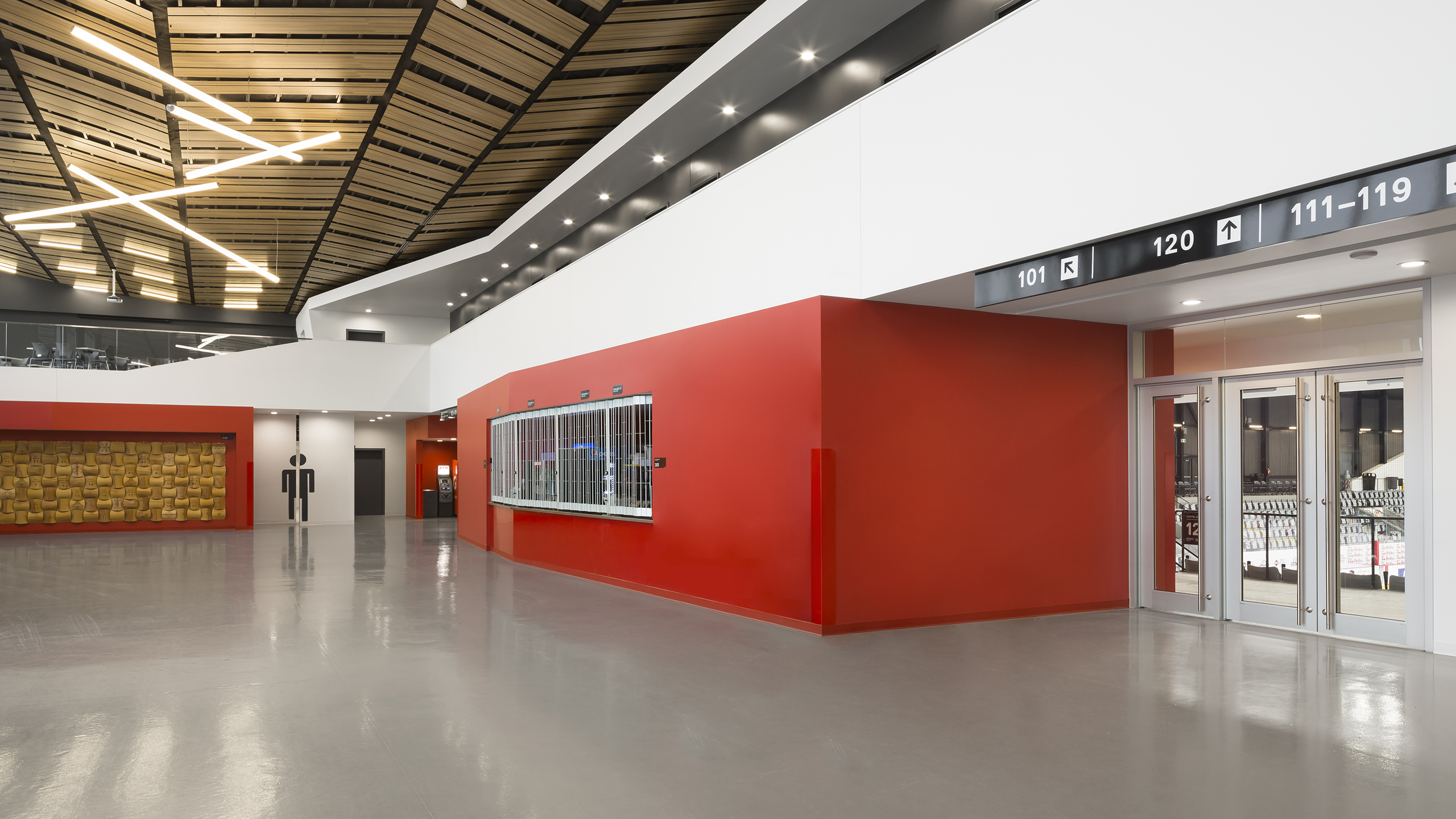Un espace public intérieur moderne et épuré avec un haut plafond, des murs rouges et blancs et des sols en béton poli. Le plafond présente des motifs géométriques en bois avec des luminaires linéaires. Les portes sur la droite indiquent les numéros de section, menant aux toilettes visibles au loin dans ce grand espace ressemblant à un amphithéâtre.