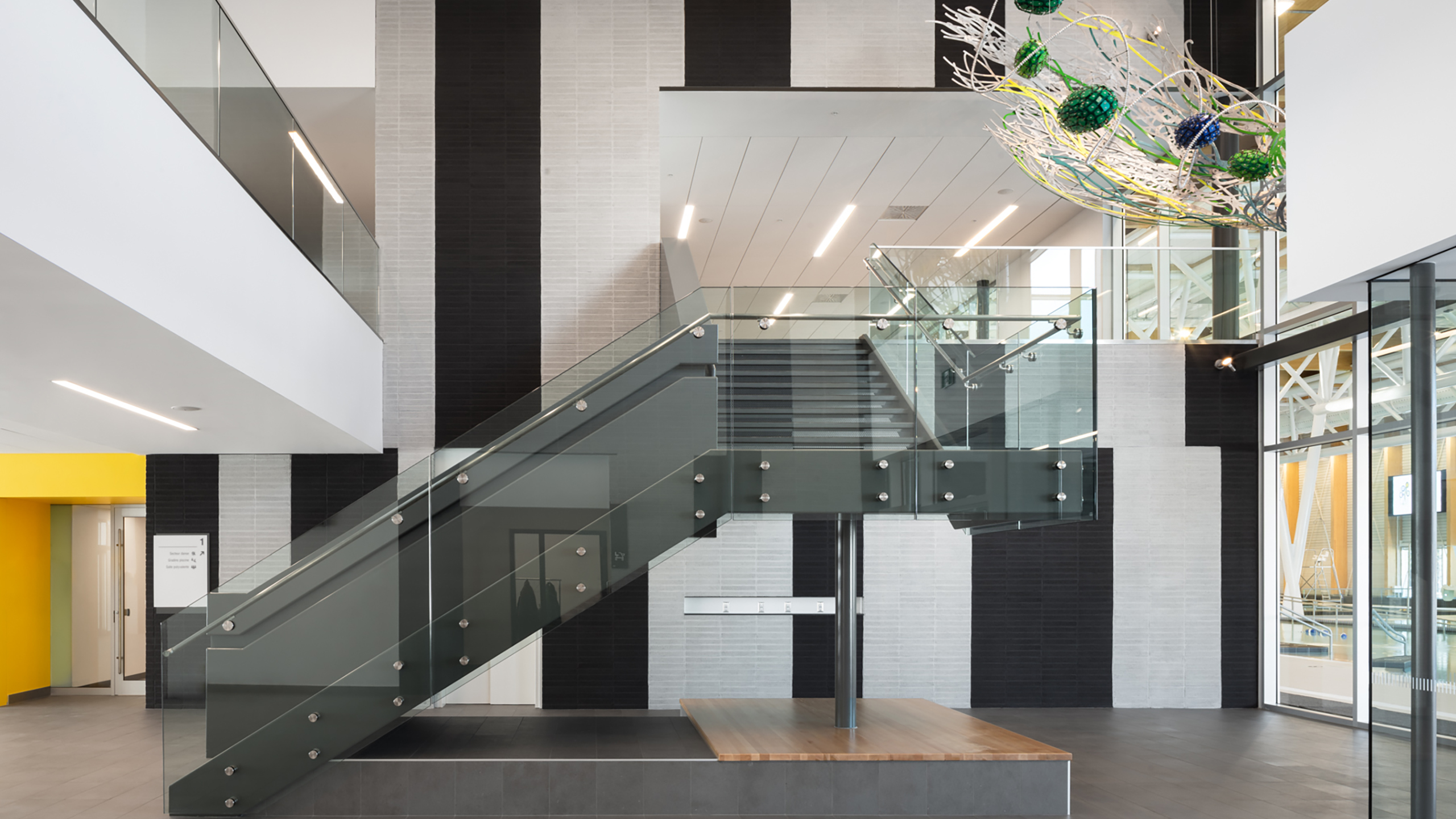 Un intérieur moderne avec un escalier en verre et en métal avec un plancher en bois à la base. La construction d'un bâtiment multifonctionnel présente un design minimaliste avec des accents noirs, blancs et jaunes. Une sculpture abstraite suspendue est suspendue au plafond. Des rampes en verre et de grandes fenêtres ajoutent à la sensation d'espace.