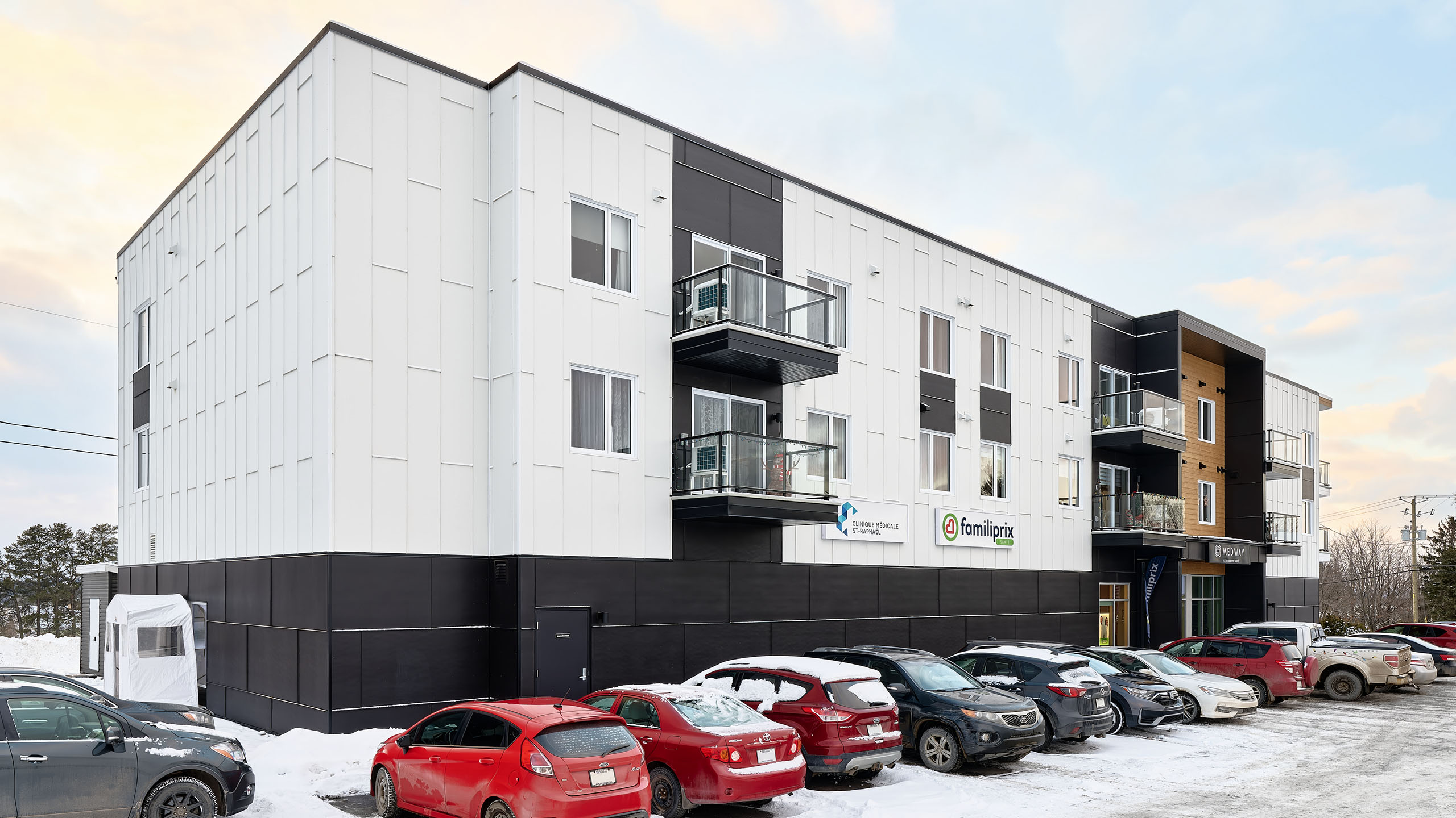 Un bâtiment moderne de trois étages aux panneaux extérieurs blancs et noirs et aux balcons aux deuxième et troisième étages marque la construction d'un nouveau bâtiment. Plusieurs voitures sont garées dans un terrain enneigé devant la structure, sur lequel on peut lire « Centracare » et « Familiprix ».