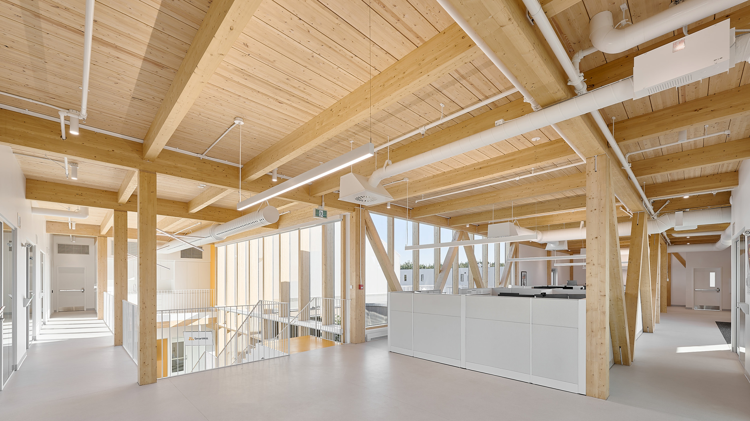 Un espace de bureau moderne aux accents de bois clair et de blanc, conçu dans le cadre de la construction d'un nouveau siège social. Des poutres en bois apparentes et de grandes fenêtres créent une atmosphère ouverte et aérée. Des bureaux et des cloisons minimalistes sont situés à droite, avec un escalier avec rampes en verre à gauche.