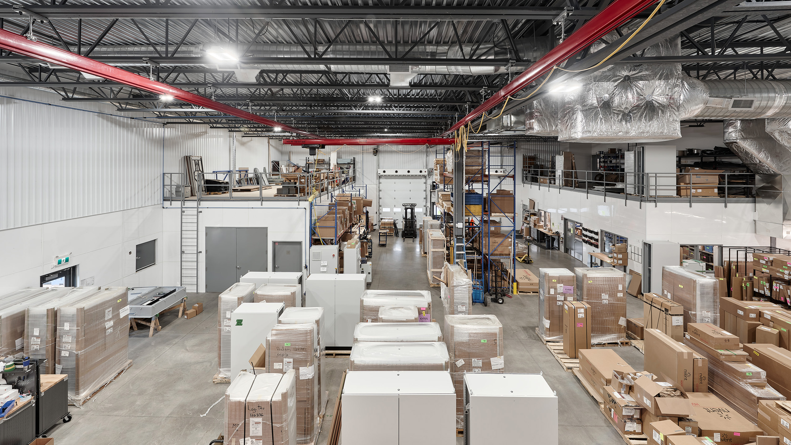 Un entrepôt spacieux avec de hauts plafonds et des étagères métalliques remplies de boîtes et de matériaux. Des palettes de marchandises sont soigneusement disposées sur le sol. Une passerelle surélevée avec un espace de stockage supplémentaire est visible sur la droite, et un éclairage industriel illumine l'espace, reflétant son rôle dans la construction d'un nouveau bâtiment.