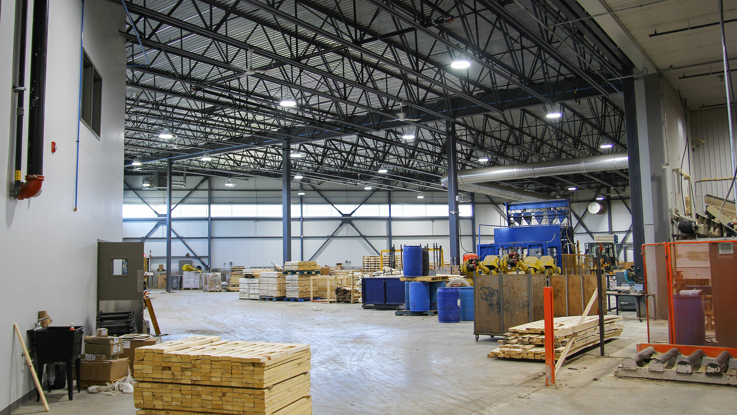 Un entrepôt industriel spacieux avec de hauts plafonds et une charpente métallique, faisant partie de l'agrandissement d'une usine existante. Des palettes en bois, des barils bleus et diverses machines sont dispersés un peu partout. Des étagères le long des murs contiennent des matériaux supplémentaires. Des plafonniers éclairent l'espace propre et organisé.
