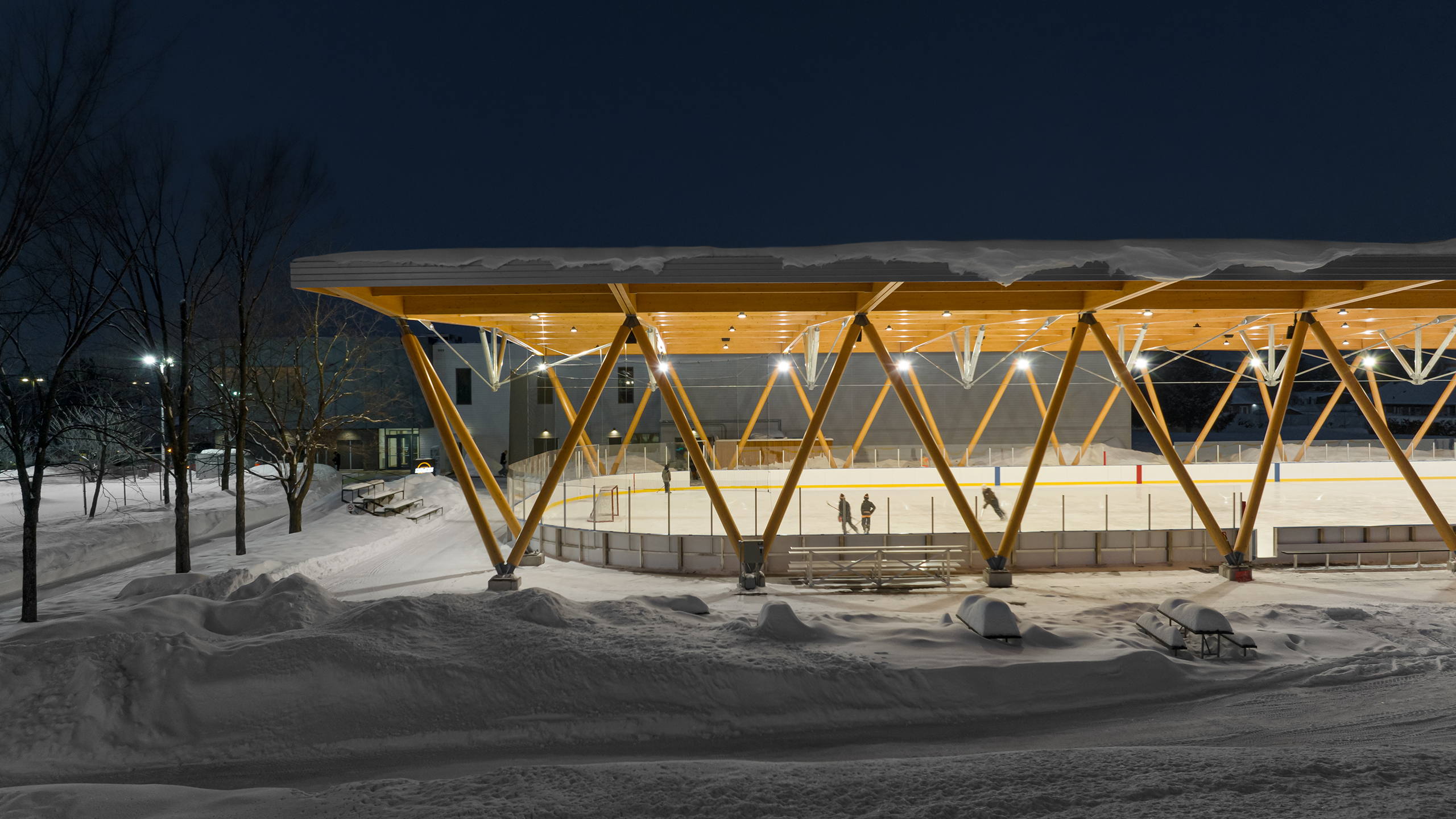 Une patinoire extérieure illuminée la nuit avec un auvent en bois moderne, illustrant une conception et une construction méticuleuses. La patinoire est entourée de terrain enneigé et d'arbres. On peut voir quelques personnes patiner sur la glace, et la zone est doucement éclairée par des lumières au plafond.