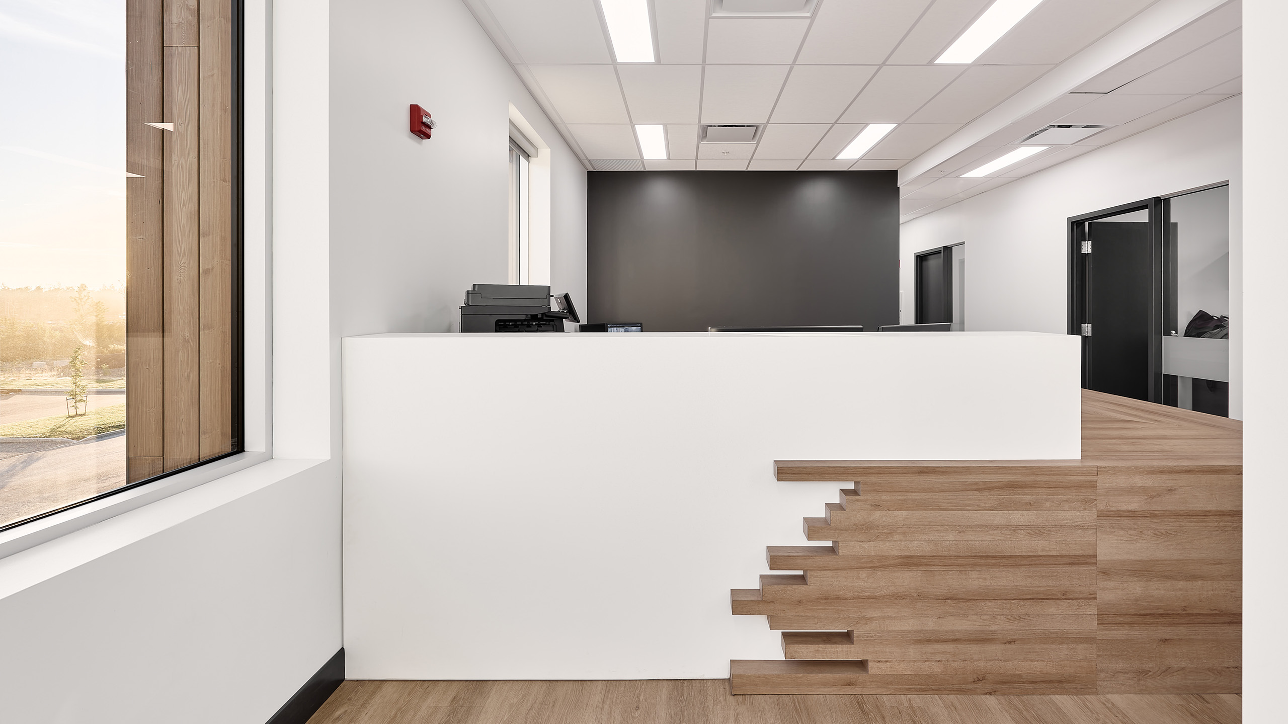 Un espace de réception de bureau moderne, faisant partie de la construction du nouveau siège social, présente un élégant bureau d'accueil blanc avec des accents en bois sur la droite. La pièce est dotée de murs blancs, d'une grande fenêtre sur la gauche et de portes menant à d'autres pièces. Un éclairage lumineux et des planchers en bois clair complètent le look.