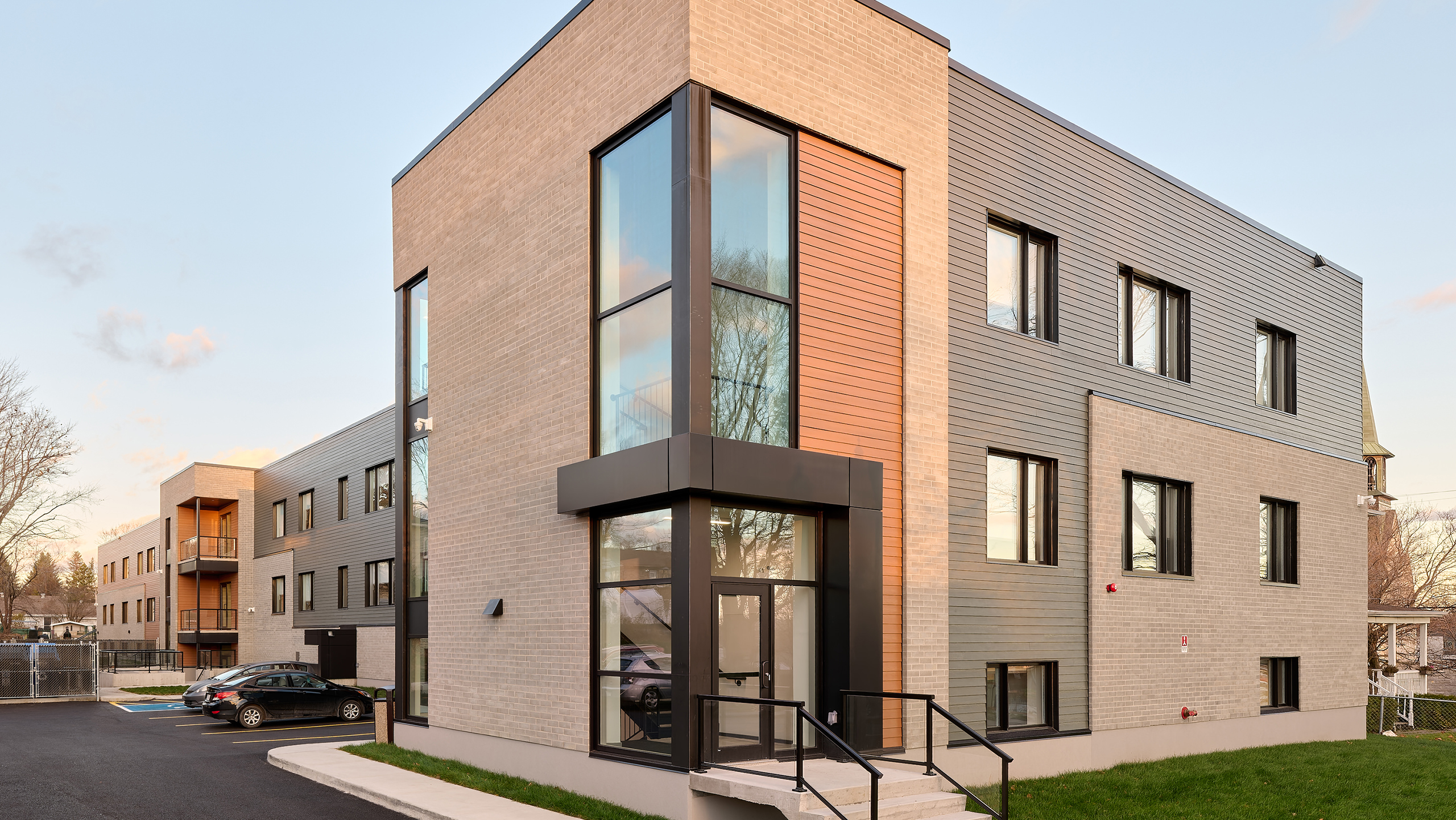 Un immeuble d'appartements moderne de trois étages avec un mélange de briques et de revêtement métallique, de grandes fenêtres en verre et un petit auvent d'entrée. Quelques voitures garées sont visibles à gauche, avec une zone herbeuse et un ciel clair en arrière-plan, mettant en valeur la récente construction d'un nouveau bâtiment.