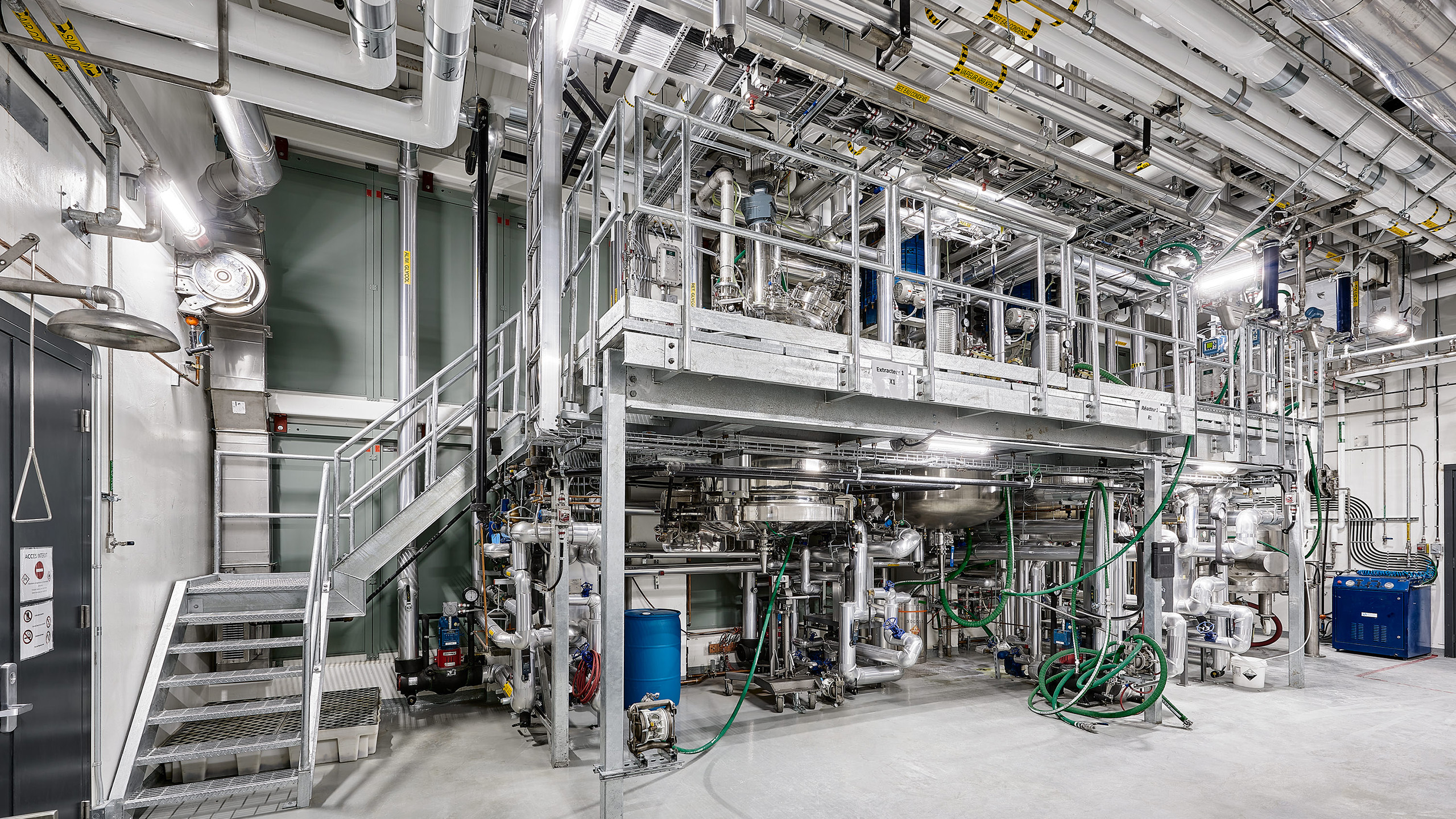 L'intérieur d'une grande installation industrielle, avec ses machines et équipements complexes, met en valeur la construction industrielle à son meilleur. La salle comprend un réseau de tuyaux, de vannes et de câbles avec des plates-formes métalliques et des escaliers pour y accéder. L'environnement bien éclairé comprend divers panneaux de contrôle et instruments visibles.