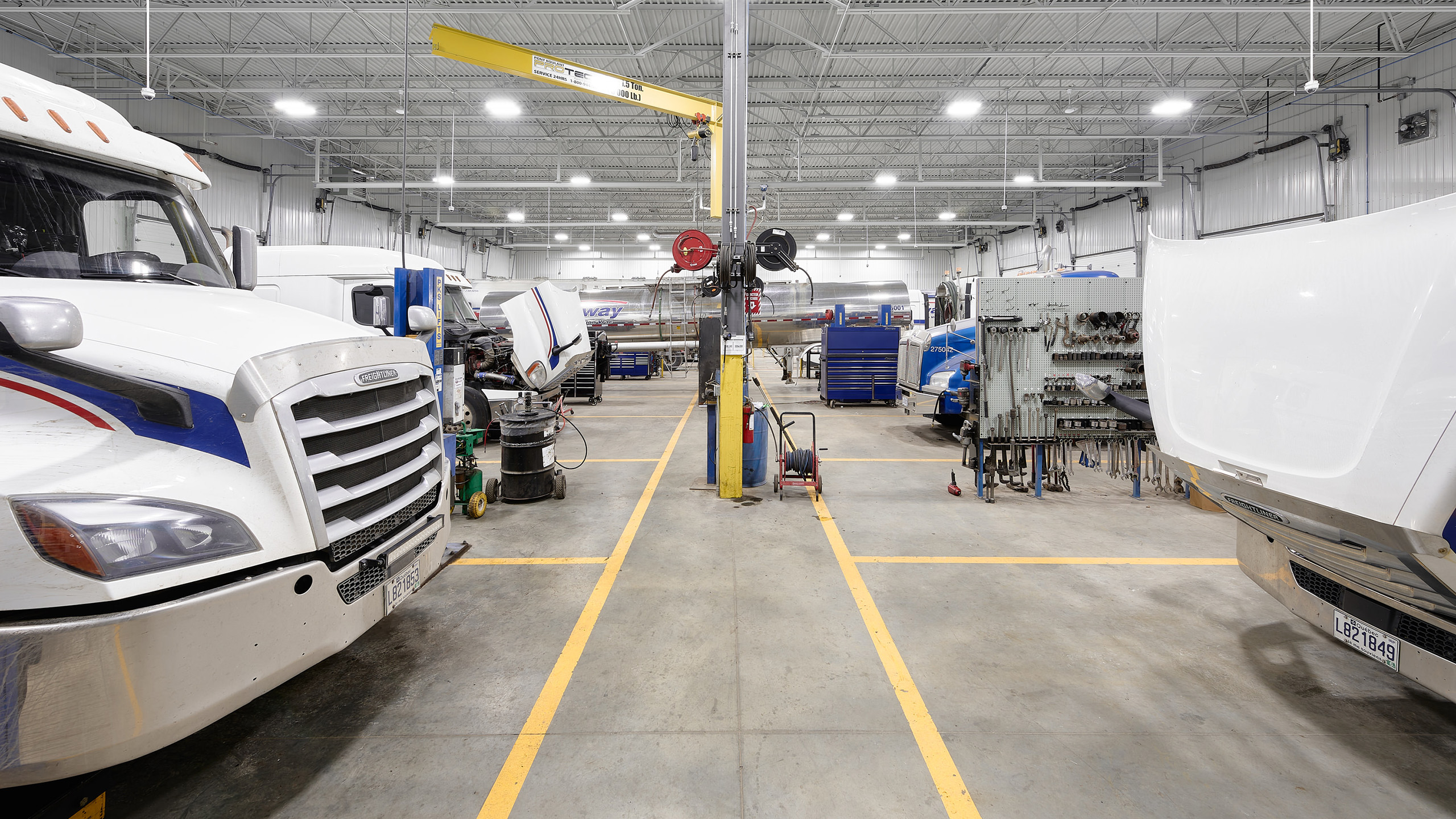 Un vaste atelier de réparation de camions avec un grand éclairage au plafond illustre la construction d'un nouveau garage. Plusieurs camions sont garés, certains avec des capots ouverts, indiquant des travaux de réparation. Divers outils et machines sont dispersés un peu partout, et un pont roulant jaune court le long du plafond. Le sol est doté de lignes de sécurité jaunes.