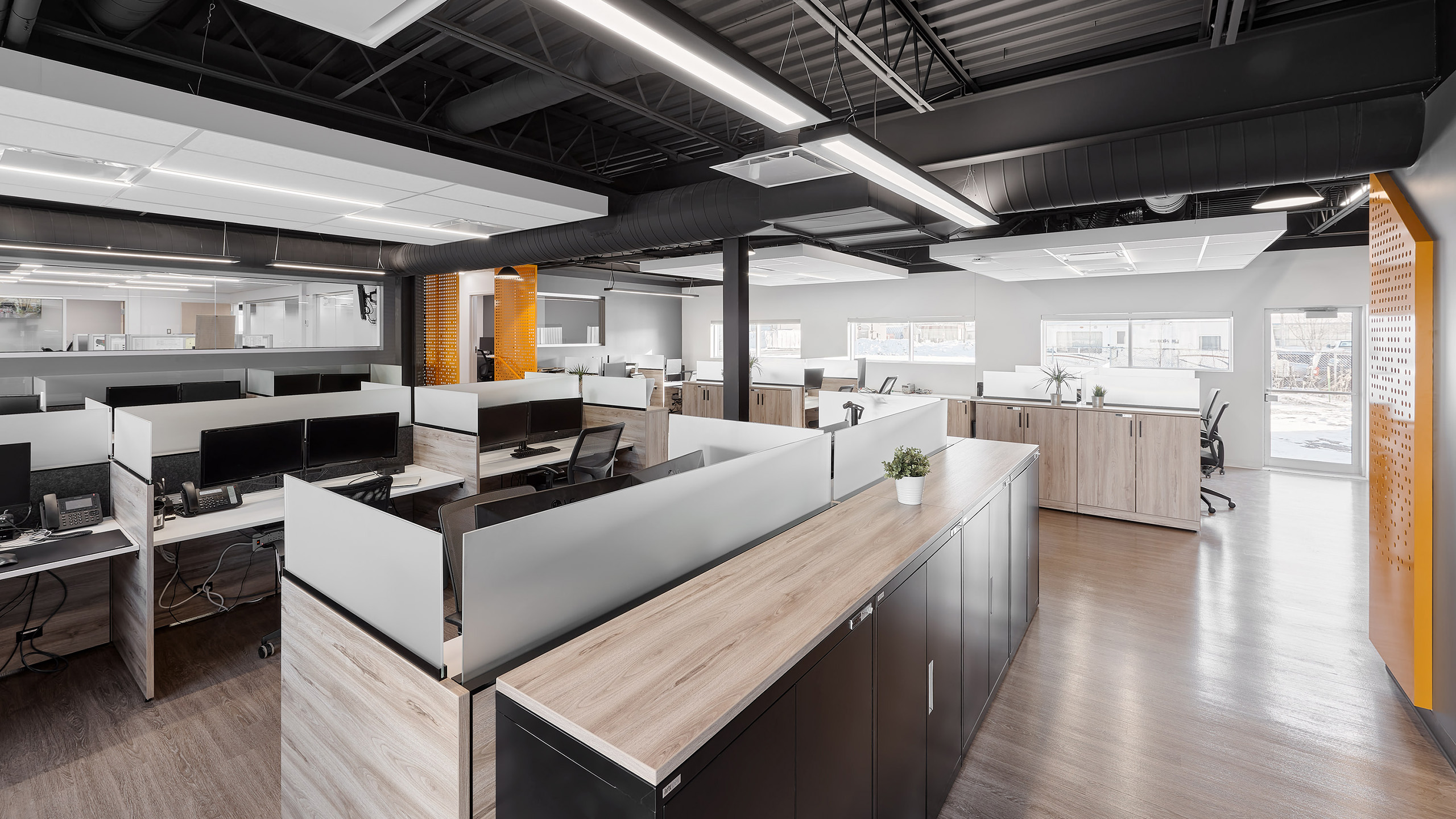 Espace de bureau moderne à aire ouverte avec plusieurs postes de travail équipés d'ordinateurs, de cloisons et de chaises ergonomiques. La récente rénovation des bureaux présente un éclairage naturel grâce à de grandes fenêtres et une décoration minimaliste avec un mélange de bois et de tons neutres.