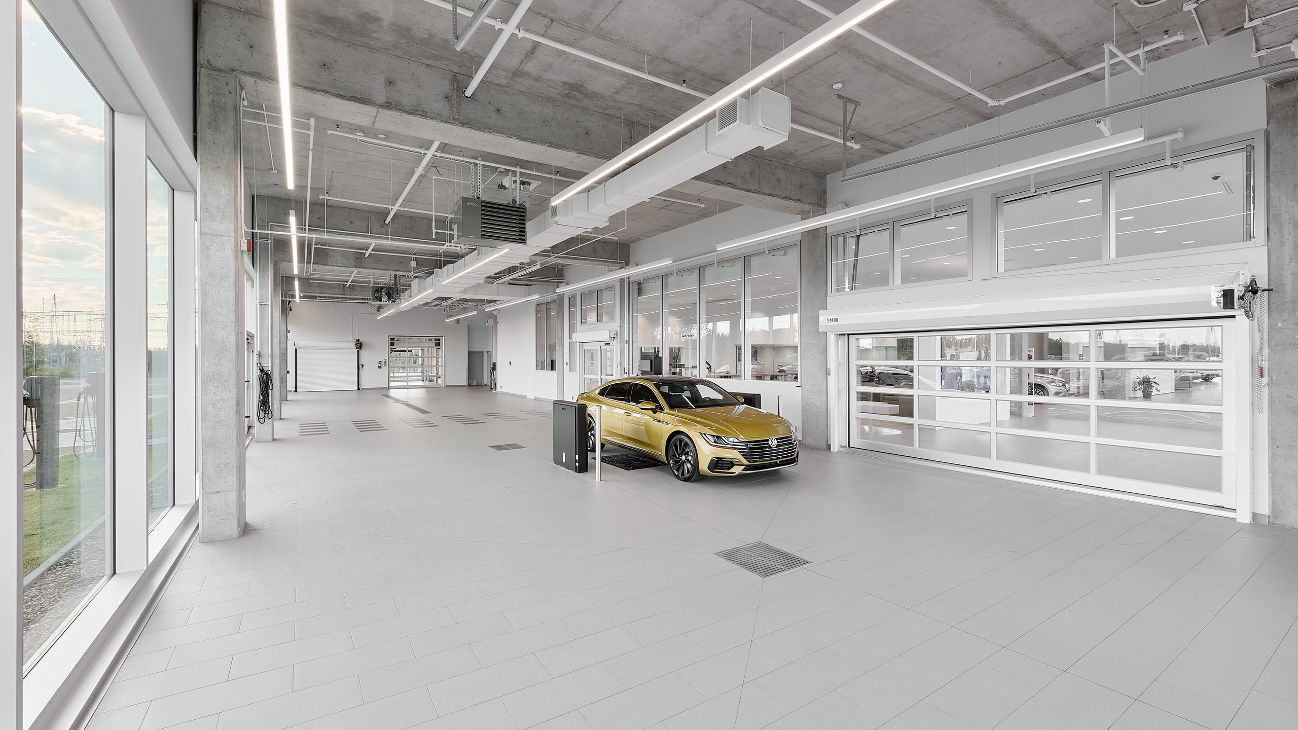 Un showroom automobile spacieux et moderne avec un haut plafond, de grandes fenêtres et des carreaux gris élégants. Une voiture dorée est garée près du centre. Des lumières fluorescentes illuminent la pièce, tandis que les portes de garage en verre fournissent une lumière naturelle abondante - l'exemple parfait de la construction d'un nouveau concessionnaire.