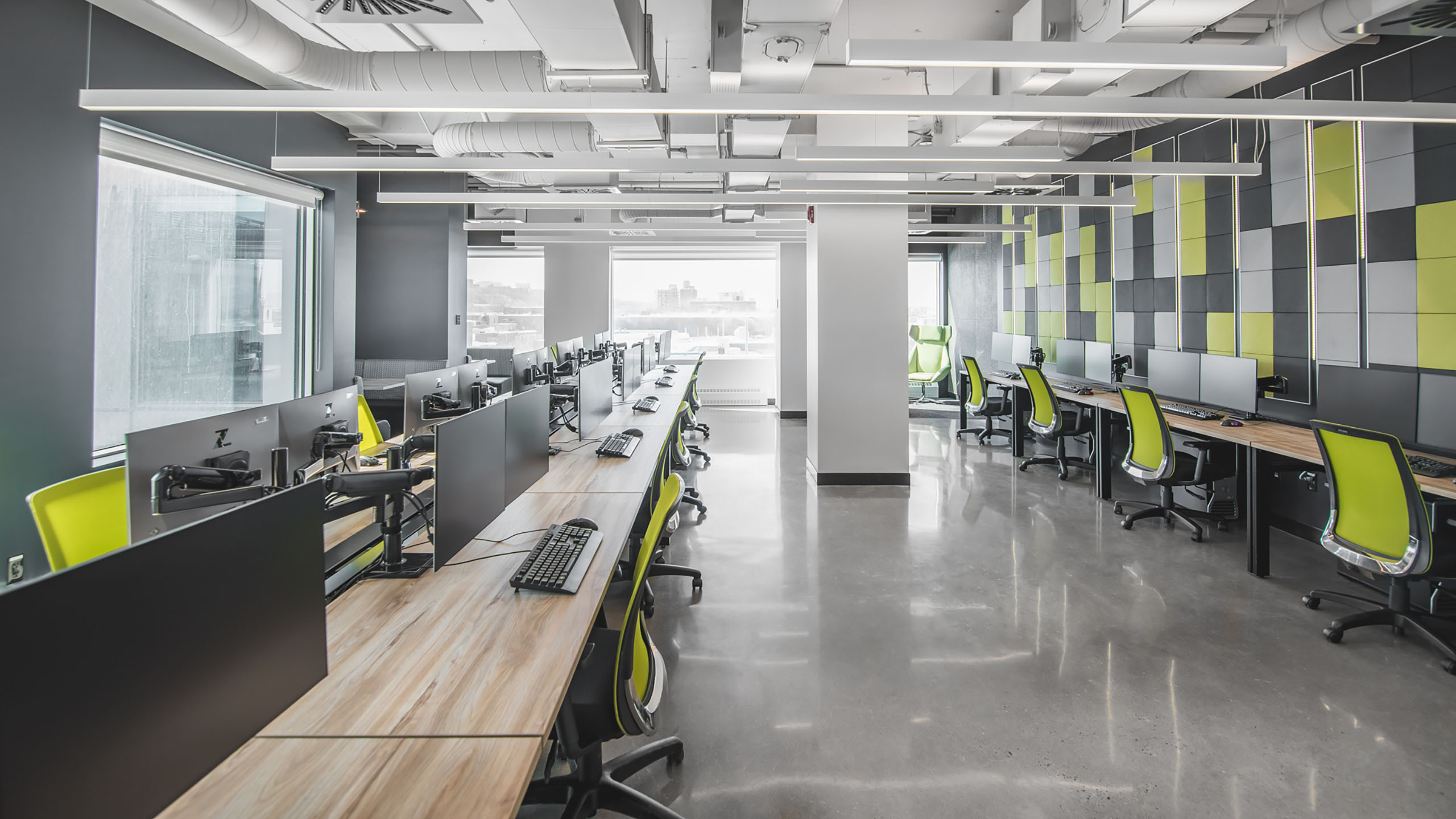 Un espace de bureau moderne et lumineux avec de grandes fenêtres comporte des rangées de bureaux équipés de deux écrans, de claviers et de chaises de bureau vertes. Les murs présentent un motif géométrique de différentes nuances de gris et de jaune. Le sol est en béton poli brillant, mettant en valeur l'impressionnante rénovation des espaces bureaux.