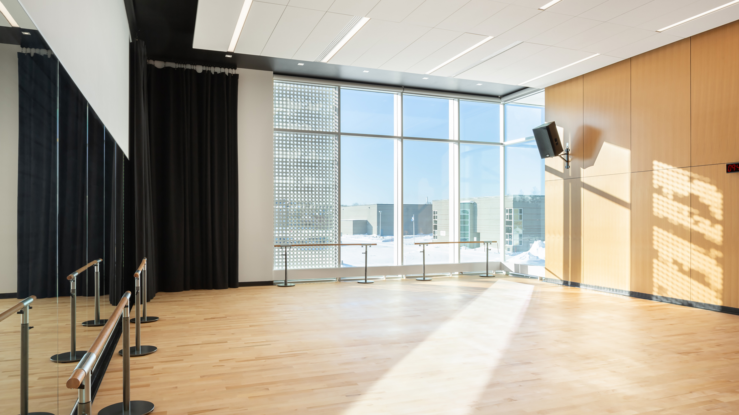 Un studio de danse spacieux et ensoleillé, doté de parquet en bois, fait partie de la construction d'un bâtiment multifonctionnel. La salle est dotée de baies vitrées d'un côté, offrant une vue dégagée sur l'extérieur. Des miroirs et des barres de ballet bordent le mur opposé. Le plafond est doté d'un éclairage encastré et une télévision est montée sur le mur de droite.
