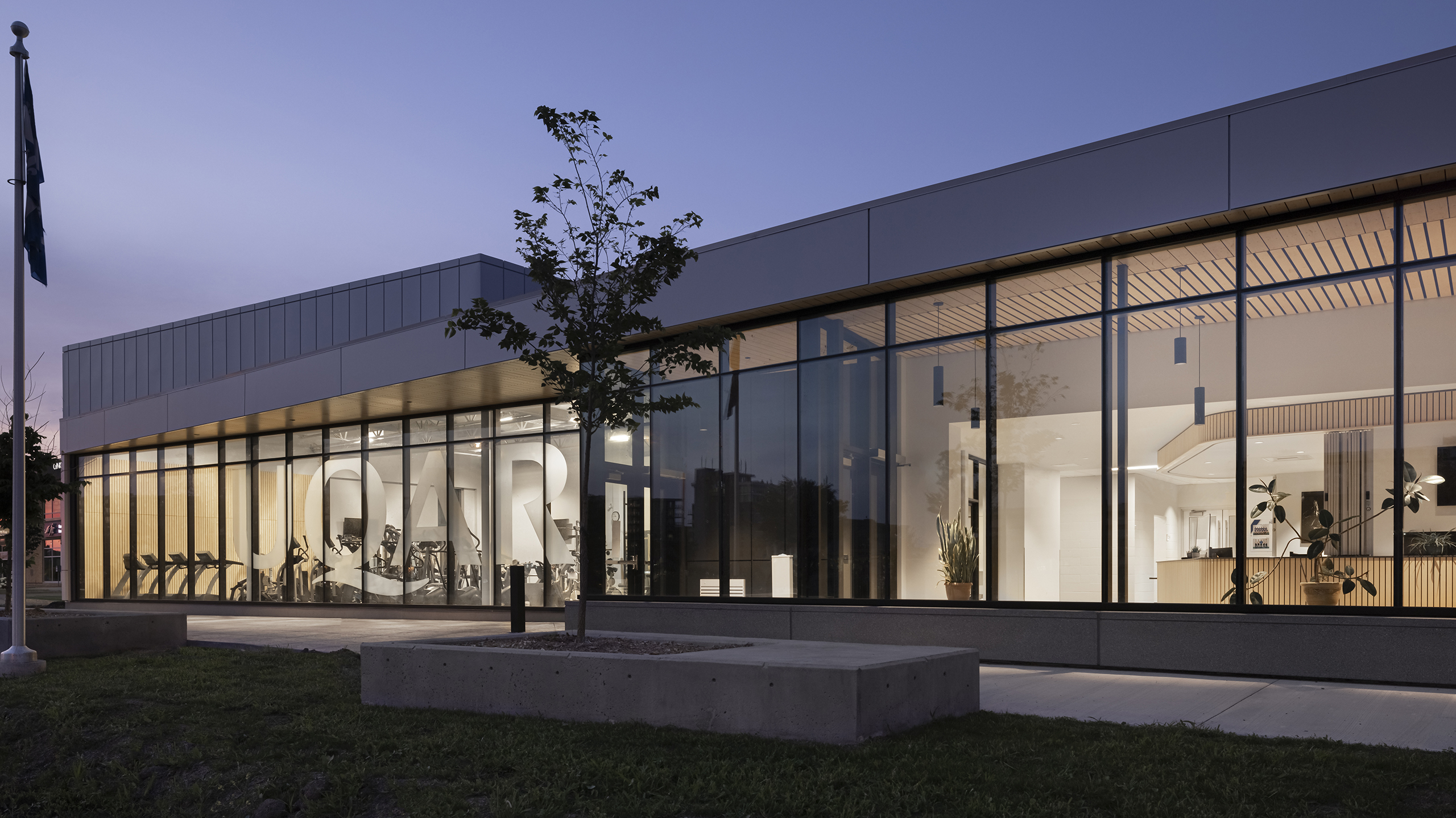 Un bâtiment moderne avec de grandes baies vitrées reflétant le ciel du soir, révélant un intérieur avec un éclairage lumineux et un design minimaliste. Un petit arbre et des blocs de béton se trouvent devant, et le drapeau est à gauche. Le mot "CAR" est visible à l'intérieur pendant la construction en cours d'un complexe sportif.