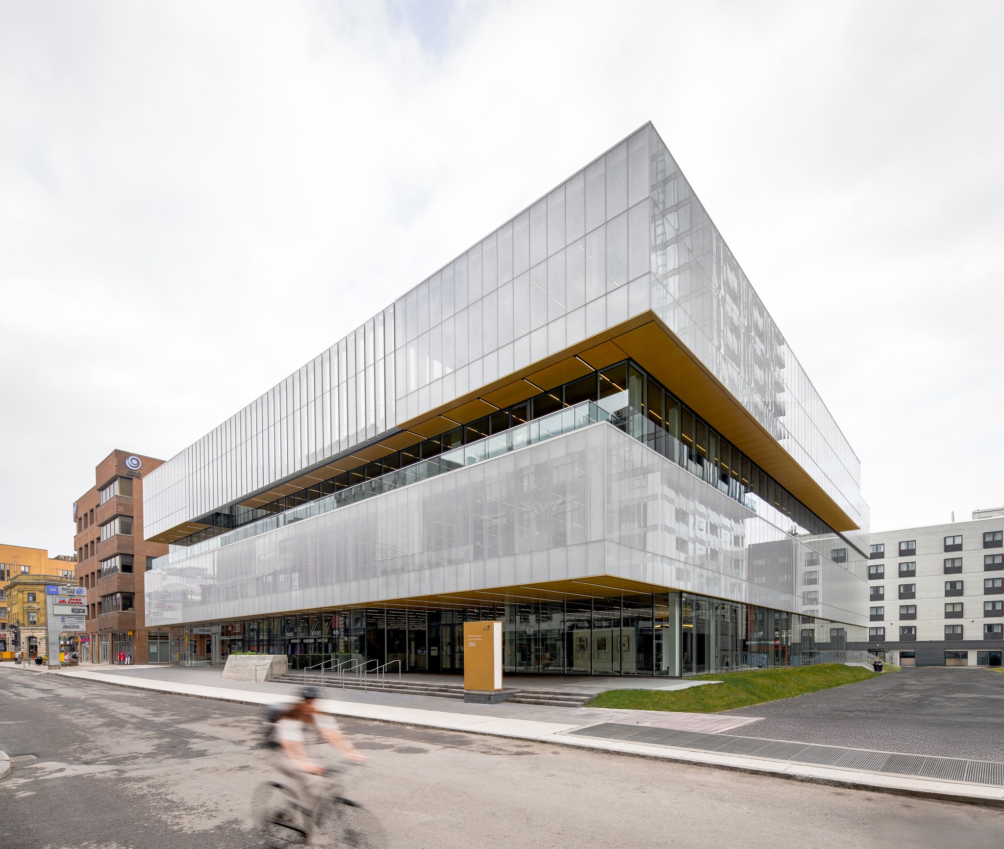 Un bâtiment moderne de plusieurs étages, avec de grands panneaux de verre et des accents jaunes, se dresse dans une rue de la ville, récemment transformée par une rénovation majeure et un agrandissement. Une personne passe à vélo, avec plusieurs autres bâtiments en arrière-plan.