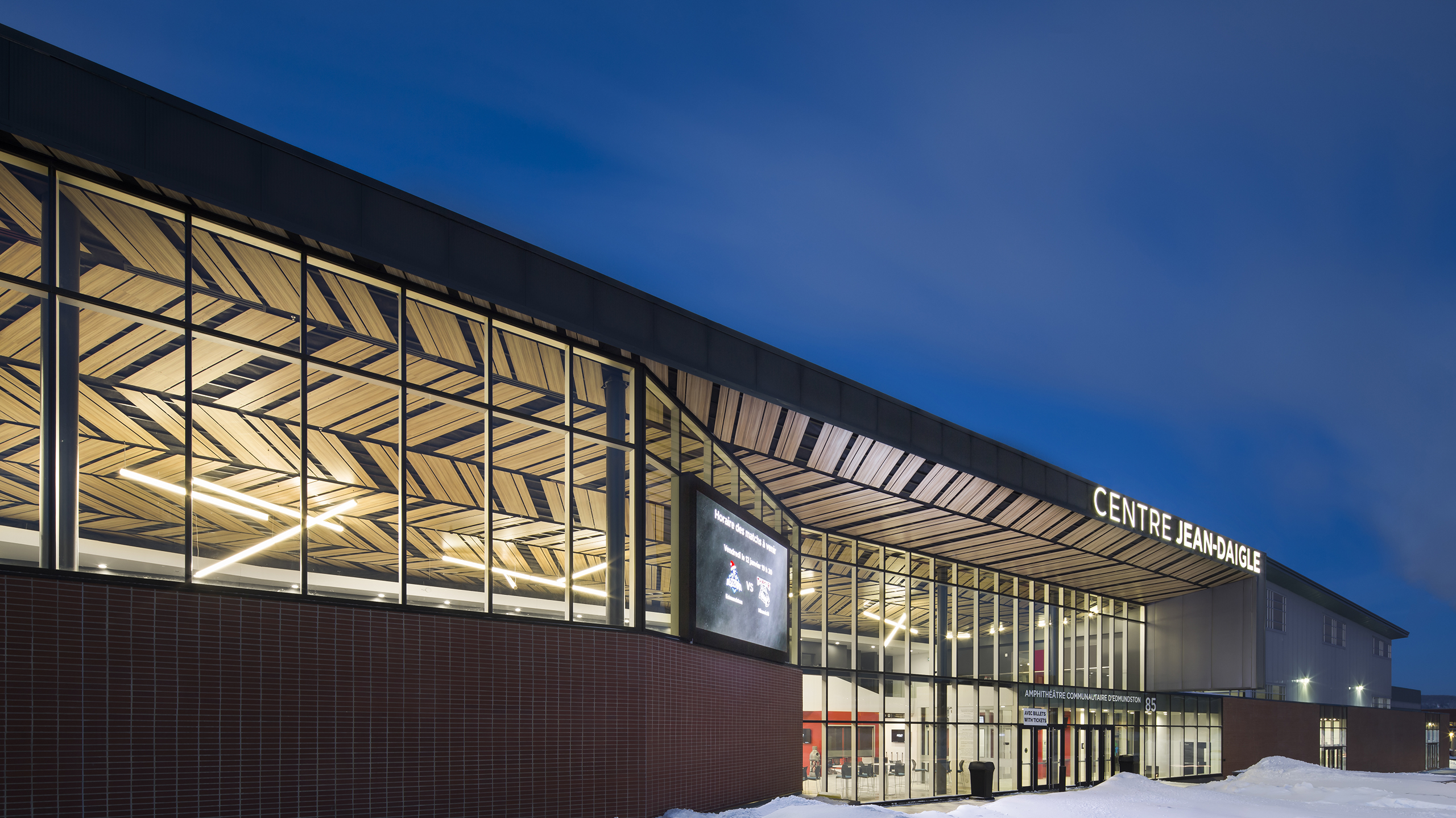 Un bâtiment moderne, baptisé « Centre Fairview », met en valeur son design contemporain avec de grandes fenêtres vitrées s'étendant sur toute la façade et un motif géométrique complexe sur le toit. La scène, rappelant un amphithéâtre de construction futuriste, se déroule le soir avec la neige recouvrant le sol au premier plan.