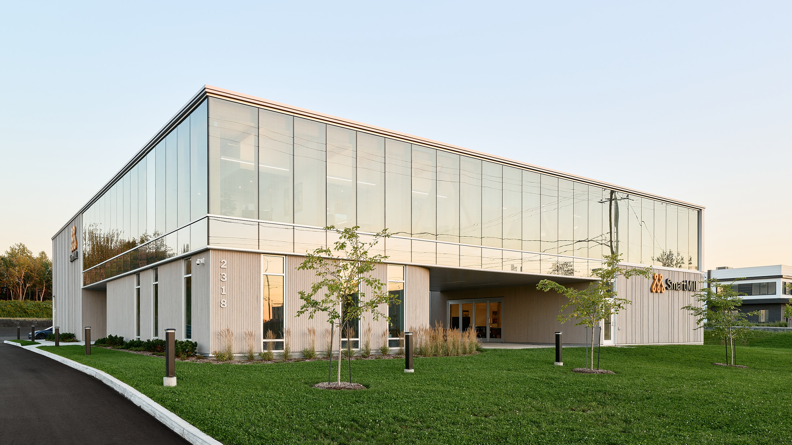 Un bâtiment moderne de deux étages avec de grandes baies vitrées reflétant l'environnement. La structure, qui fait partie de la construction d'un nouveau siège social, présente une conception rectangulaire avec un surplomb couvrant l'entrée. Il est situé sur un terrain gazonné bien entretenu avec quelques jeunes arbres et des lampadaires.