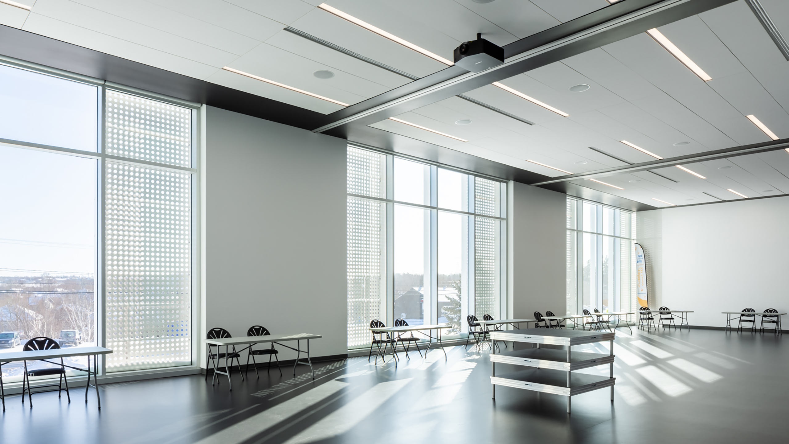 Une grande salle de conférence moderne avec des fenêtres allant du sol au plafond laissant entrer une abondante lumière naturelle, cet espace illustre la vision derrière la construction d'un bâtiment multifonctionnel. La salle comprend un haut plafond, un rétroprojecteur, plusieurs tables et chaises, des murs blancs et un sol noir.