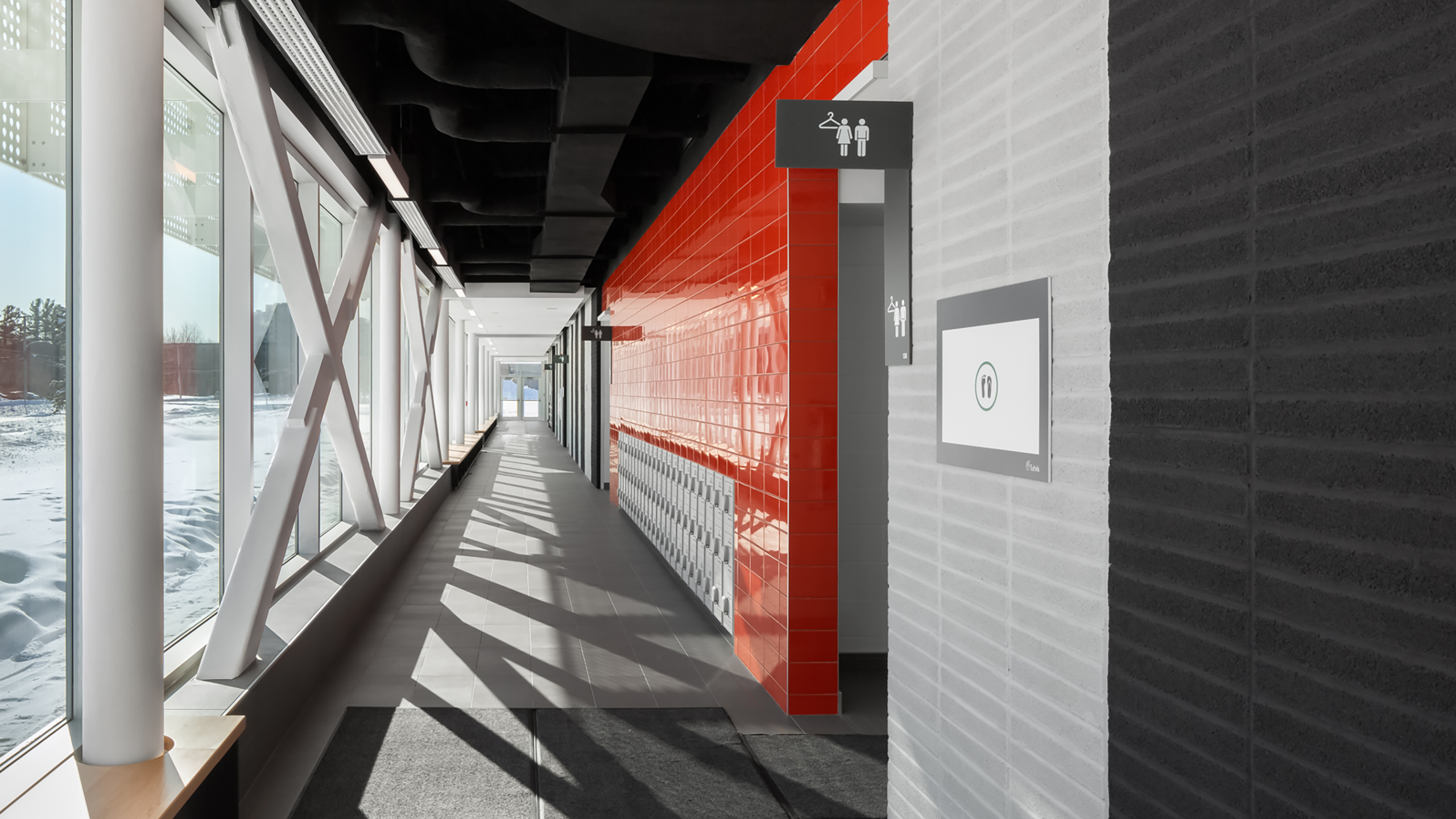 Un couloir moderne avec de grandes fenêtres à gauche, laissant entrer la lumière naturelle. Le côté droit présente un mur carrelé rouge avec des panneaux indiquant les toilettes. Le couloir a un sol gris et un banc de couleur claire. De la neige est visible à l'extérieur des fenêtres, mettant en valeur la construction d'un bâtiment multifonctionnel.