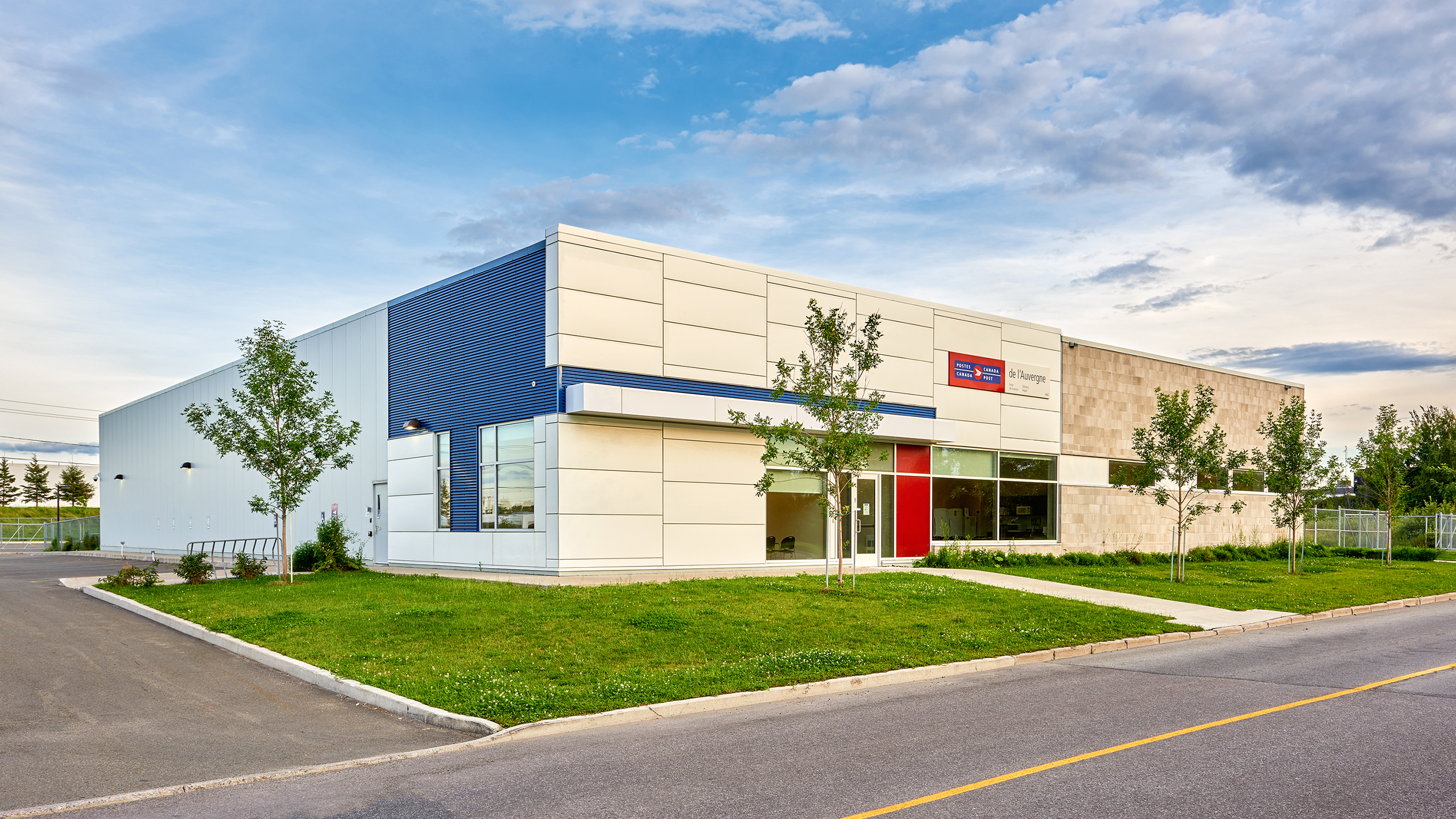 Un bâtiment commercial moderne bicolore aux murs extérieurs blancs et bleus se dresse sur un terrain d'angle. Ce centre de construction est doté de grandes fenêtres et d'une porte d'entrée rouge. Le paysage est bien entretenu avec de jeunes arbres et de l'herbe verte, sous un ciel partiellement nuageux.