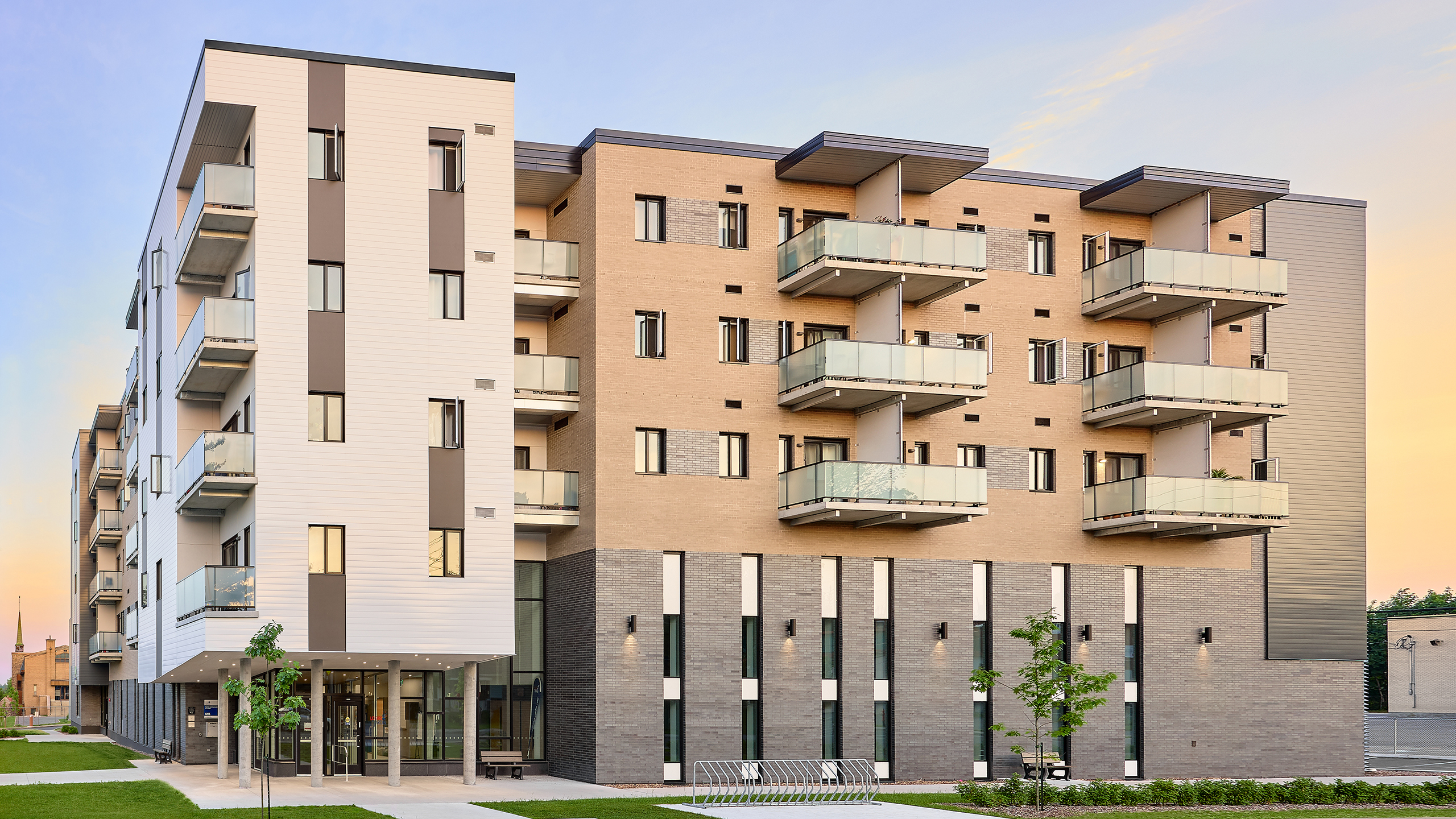 Un immeuble multirésidentiel moderne de cinq étages avec un mélange de briques et de revêtement extérieur blanc. Chaque unité dispose d'un balcon avec garde-corps en verre. Le rez-de-chaussée comprend une entrée avec de grandes fenêtres. Il y a quelques arbres et un banc dans l'espace paysager en face.