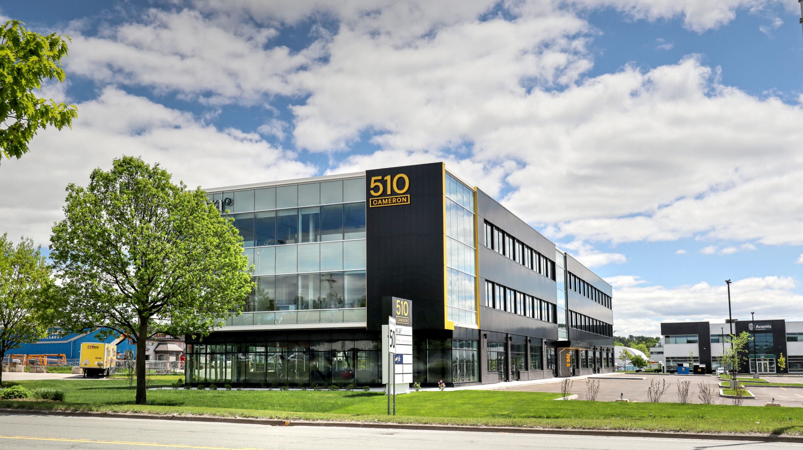 Un immeuble de bureaux moderne de trois étages avec de grandes baies vitrées, un panneau indiquant "510 Carrefours" à l'angle, et des arbres à proximité - un exemple impressionnant de construction d'un bâtiment commercial sous un ciel partiellement nuageux.