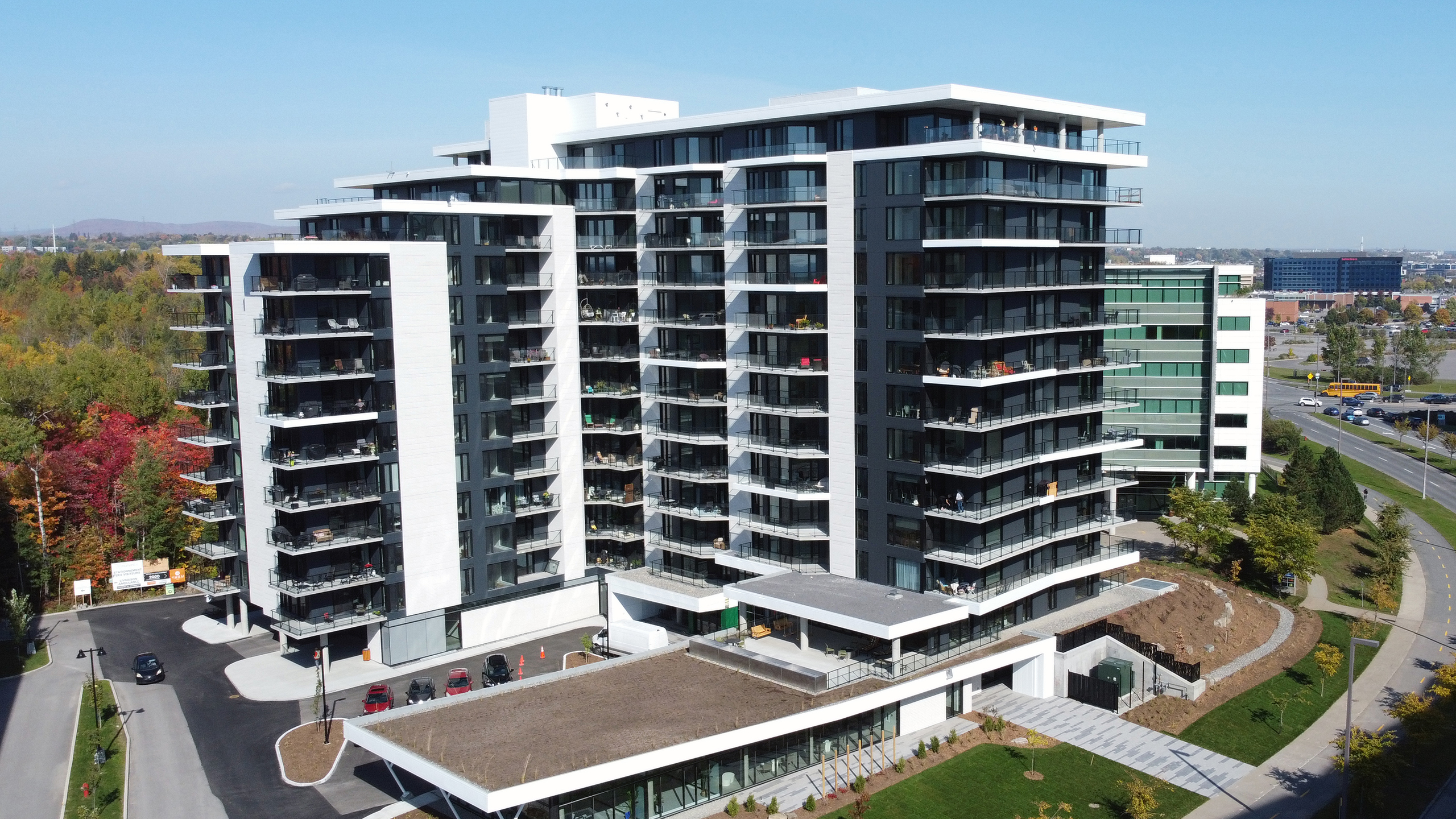 Un immeuble multirésidentiel moderne, doté de multiples balcons et d'un toit plat, situé au milieu d'un espace paysager. Entouré de verdure, d'arbres, d'un petit parking et de routes à proximité. D'autres bâtiments sont visibles en arrière-plan.
