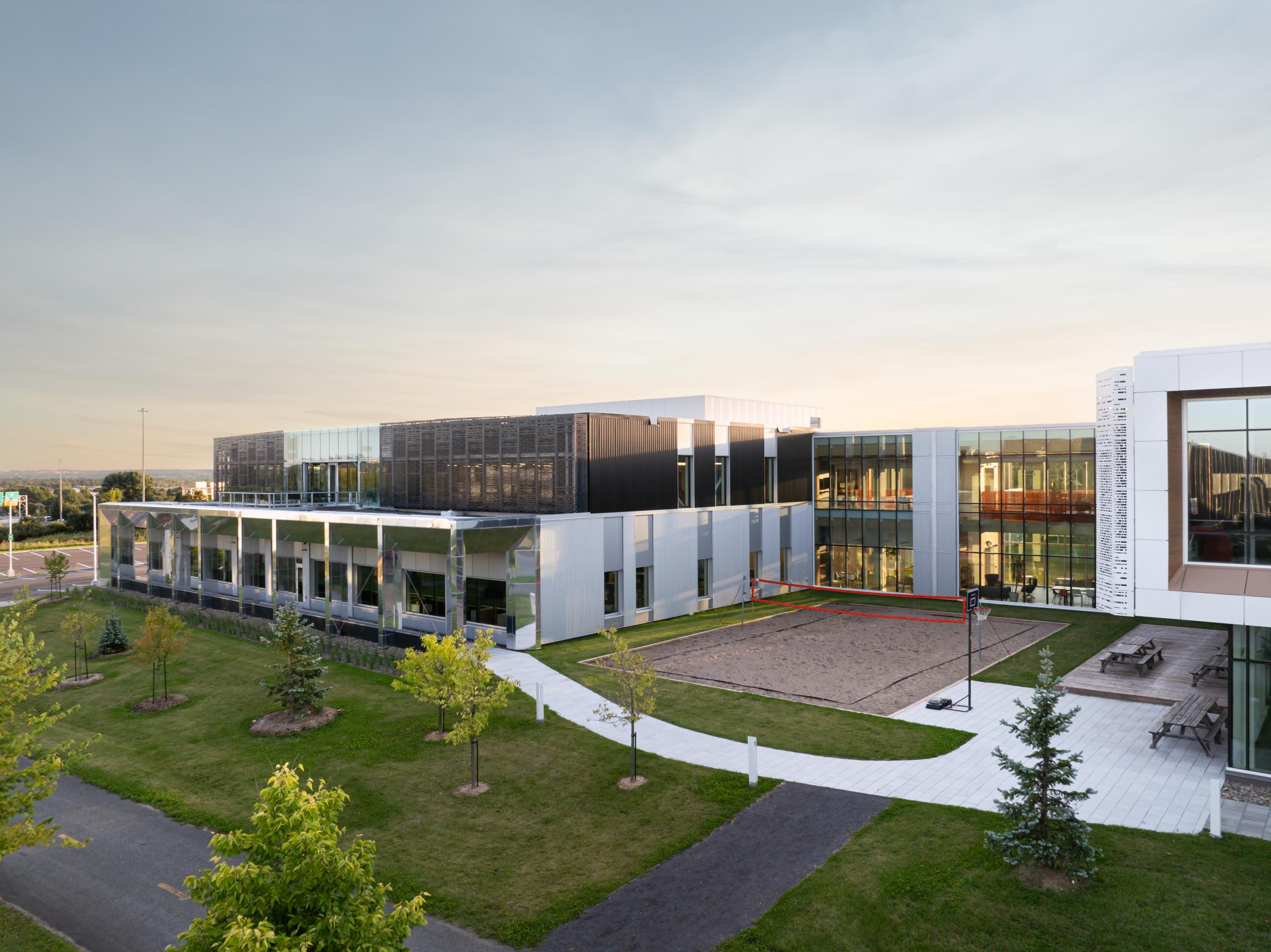 Un bâtiment moderne de plusieurs étages avec de grandes baies vitrées, entouré de pelouses vertes, d'arbres, de sentiers de promenade et d'un terrain de volley-ball sur sable dans la cour, sous un ciel nuageux au coucher du soleil.