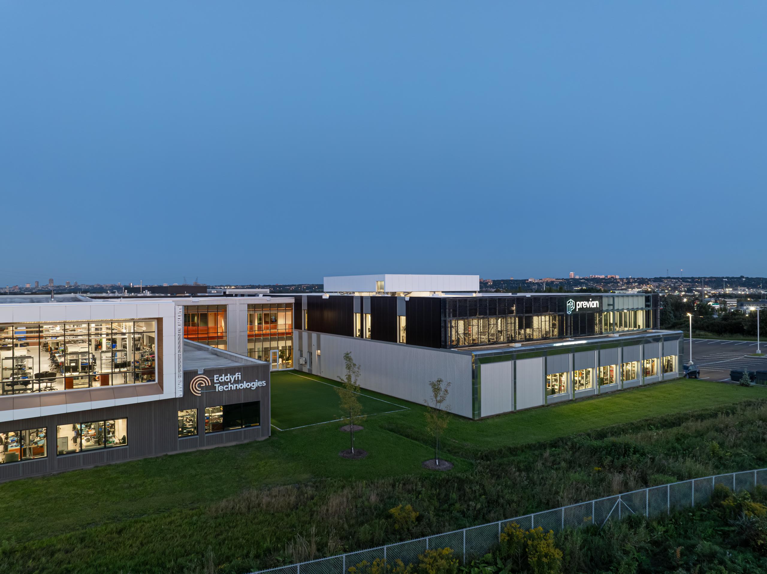 Un immeuble de bureaux moderne et un centre technologique au crépuscule, avec les logos "Eddyfi Technologies" et "Previan" à l'extérieur, entouré d'herbe verte et d'une clôture, la ligne d'horizon de la ville étant faiblement visible en arrière-plan.