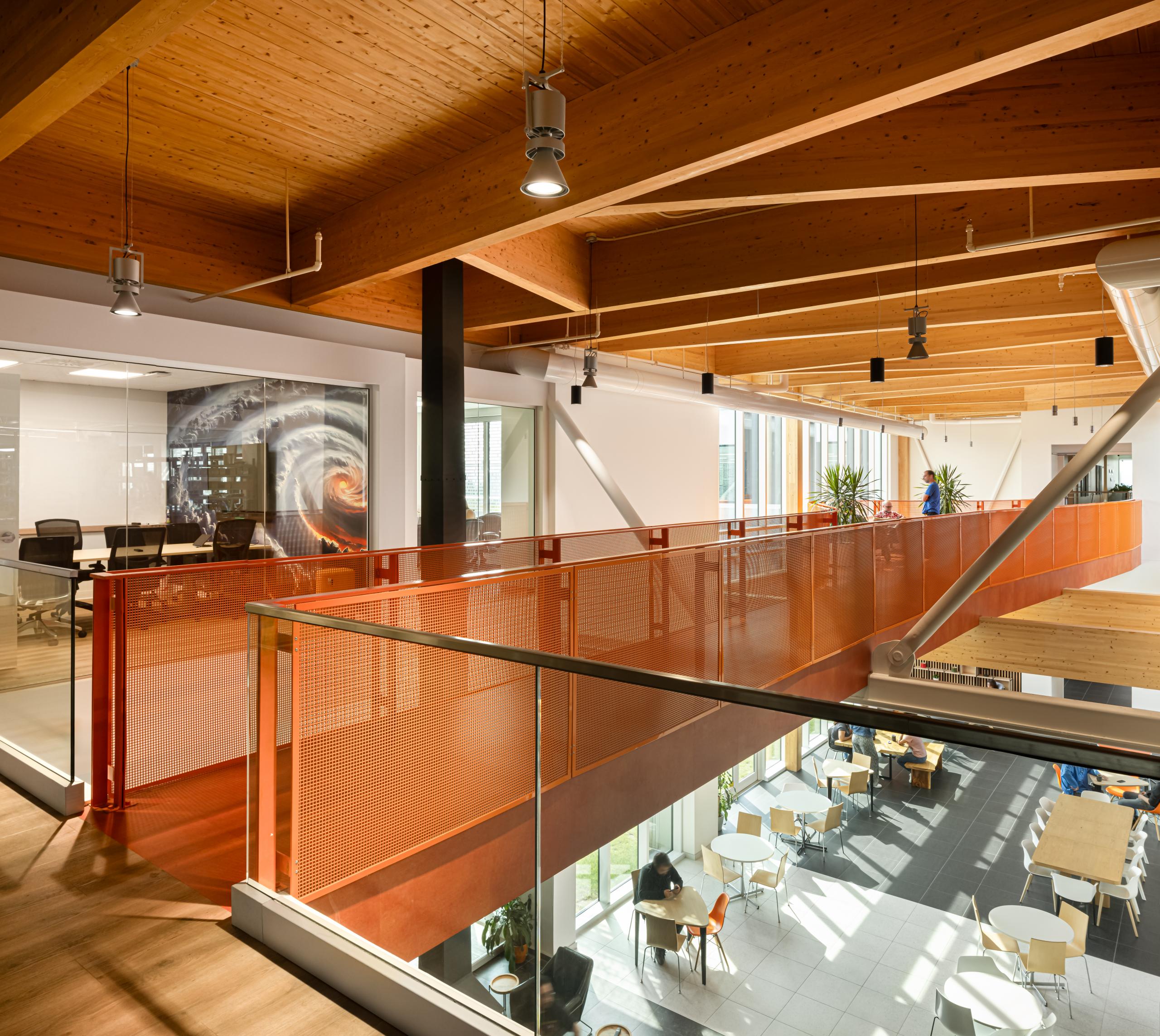 Intérieur de bureau moderne avec des plafonds en bois, des garde-corps en métal orange, des salles de réunion vitrées et un atrium ouvert avec des tables, des chaises et la lumière naturelle des grandes fenêtres.