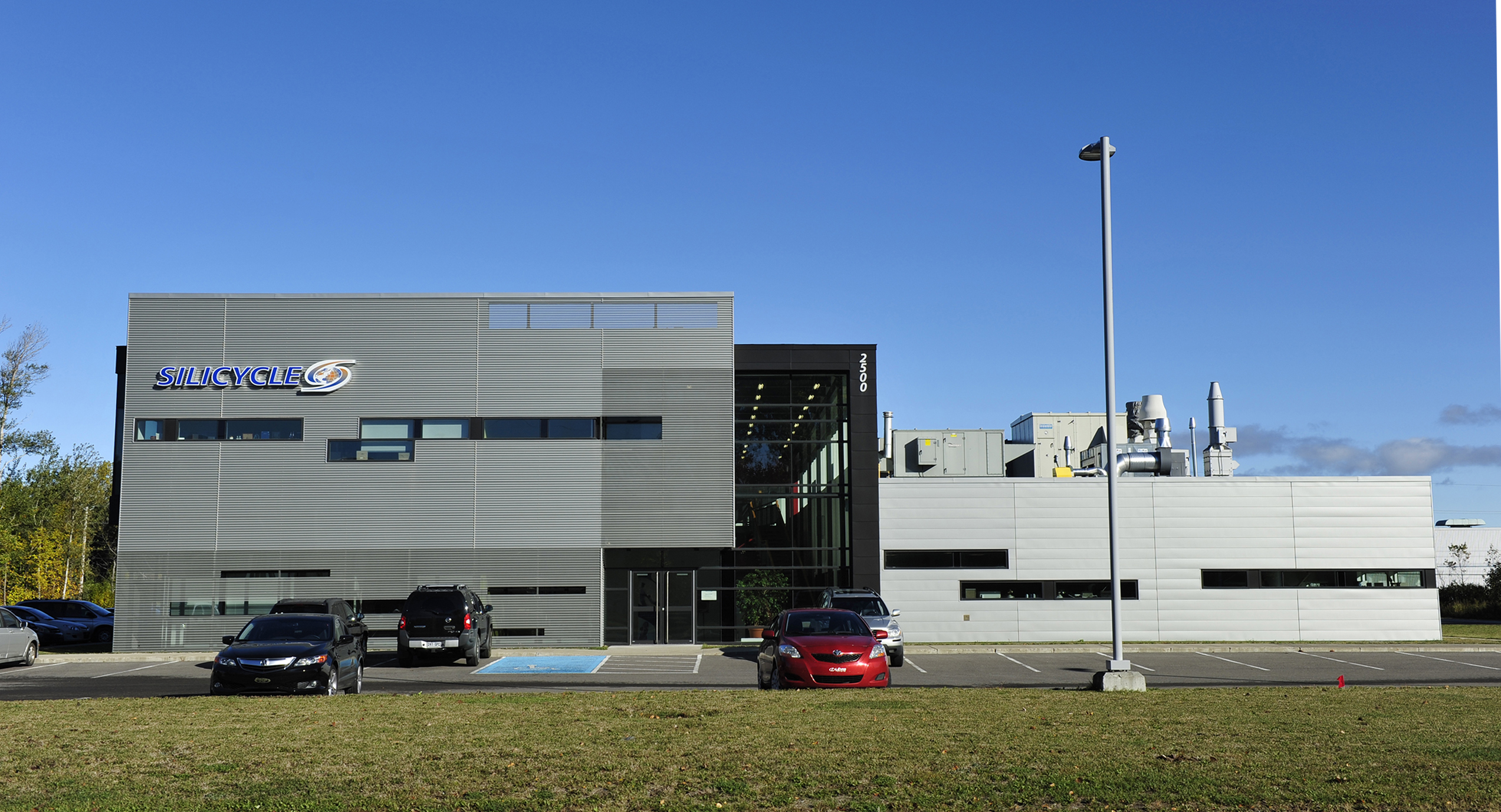 Un bâtiment moderne, rectangulaire, avec le logo « Silicycle » dans le coin supérieur gauche. Le bâtiment est gris avec de grandes fenêtres et un parking devant, contenant plusieurs voitures. Un grand lampadaire est visible dans le parking, se détachant sur un ciel bleu clair.