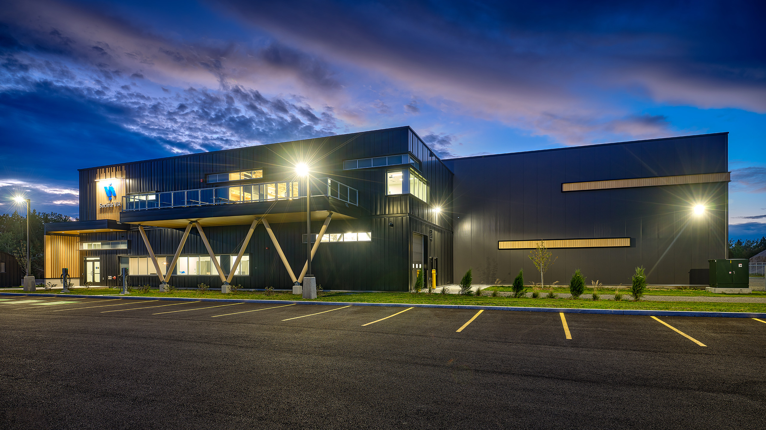 Un bâtiment moderne de deux étages avec de grandes fenêtres en verre et des panneaux métalliques élégants est illuminé au crépuscule. Il y a plusieurs lumières sur le bâtiment et quelques petits arbres sont plantés devant. Le parking vide au premier plan est orné de lignes jaunes soigneusement peintes pour les places de stationnement, faisant allusion à la construction d'une nouvelle usine.