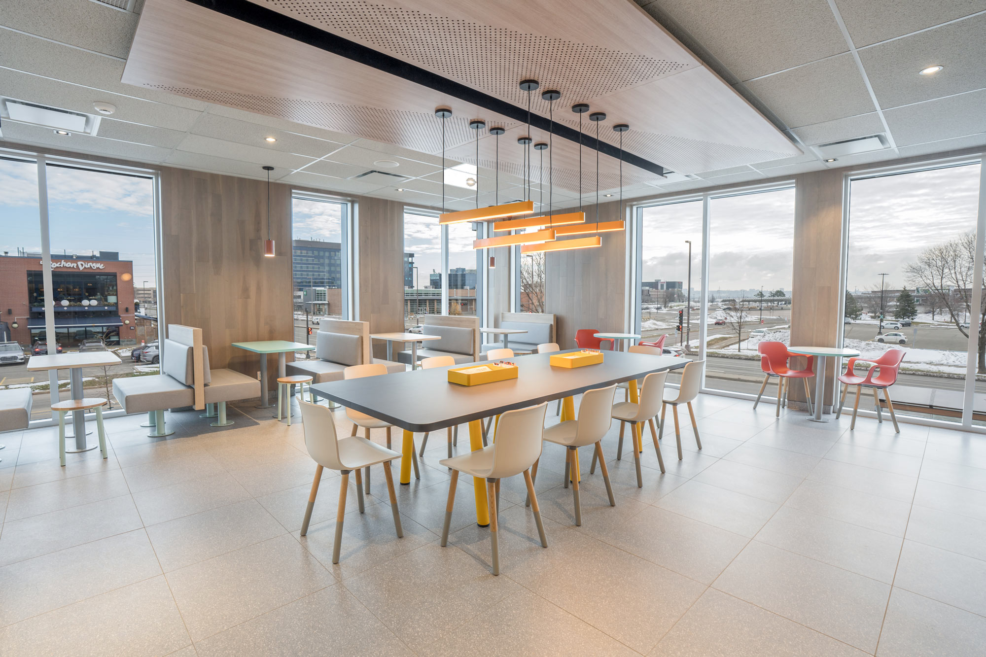 A modern, bright dining area with a long communal table, individual tables, white and gray chairs, large windows, pendant lights, and views of buildings and a snowy landscape outside.