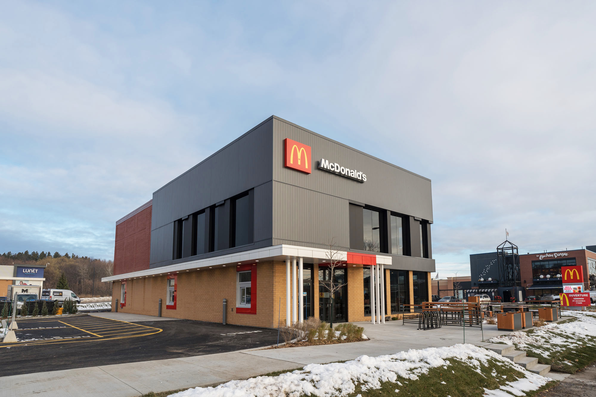 A modern McDonald's restaurant, recently completed as part of a construction d’un restaurant project, features large windows and outdoor seating amidst patches of snow under a partly cloudy sky. Another McDonald's sign appears on a nearby building.