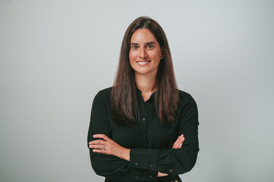 Une femme aux longs cheveux bruns, vêtue d'une chemise noire à boutons, se tient sur un fond gris clair uni, les bras croisés, et sourit à l'appareil photo.
