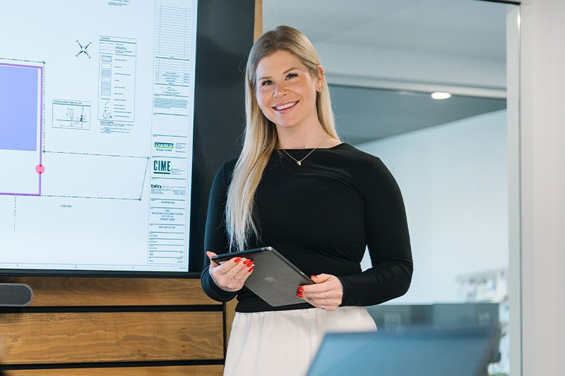 Une femme aux longs cheveux blonds, vêtue d'un haut noir et d'un pantalon blanc, se tient devant un écran affichant des diagrammes techniques, souriante et tenant une tablette.